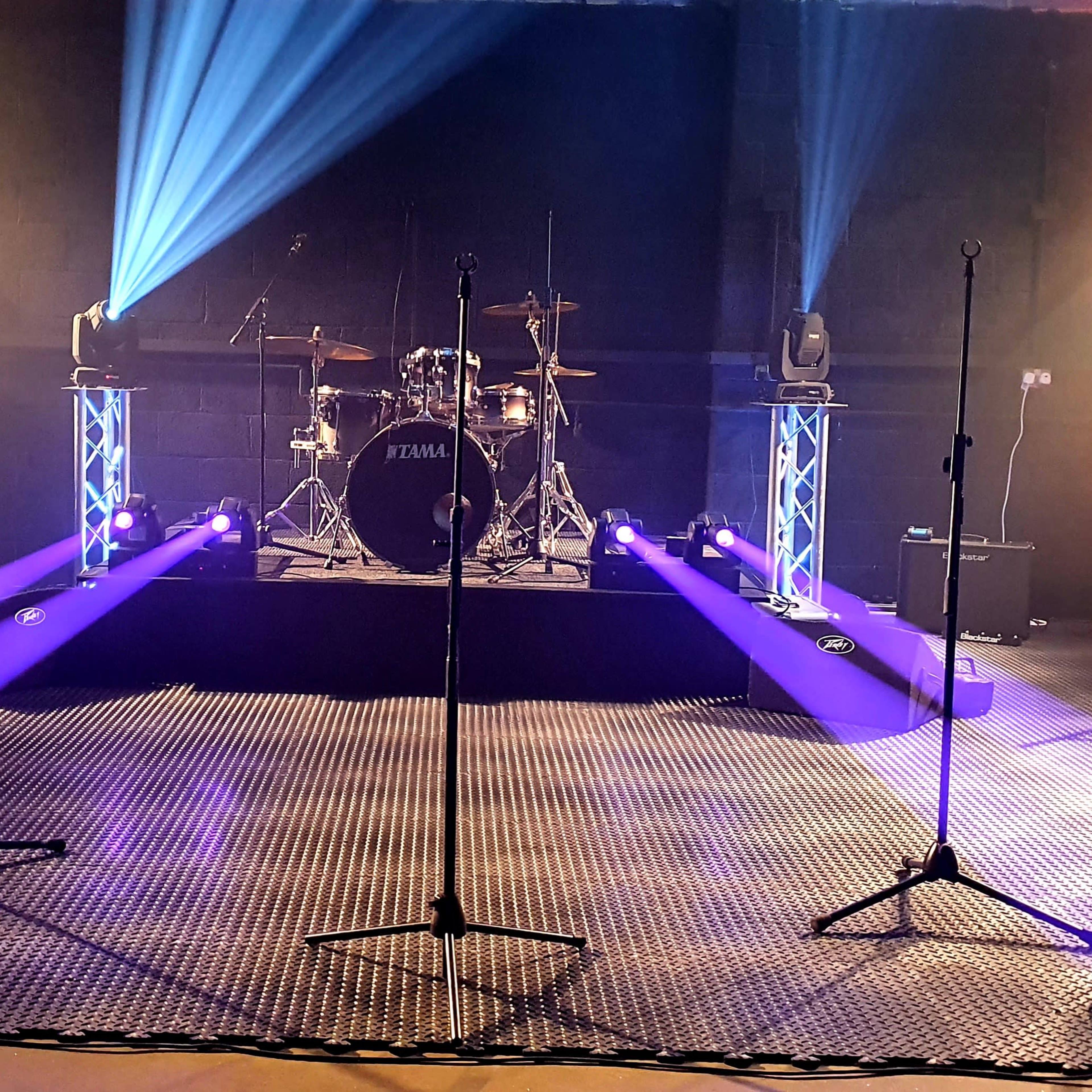 A stage setup for a concert, featuring a drum set at the center, multiple microphones on stands, and colored lights illuminating the area.