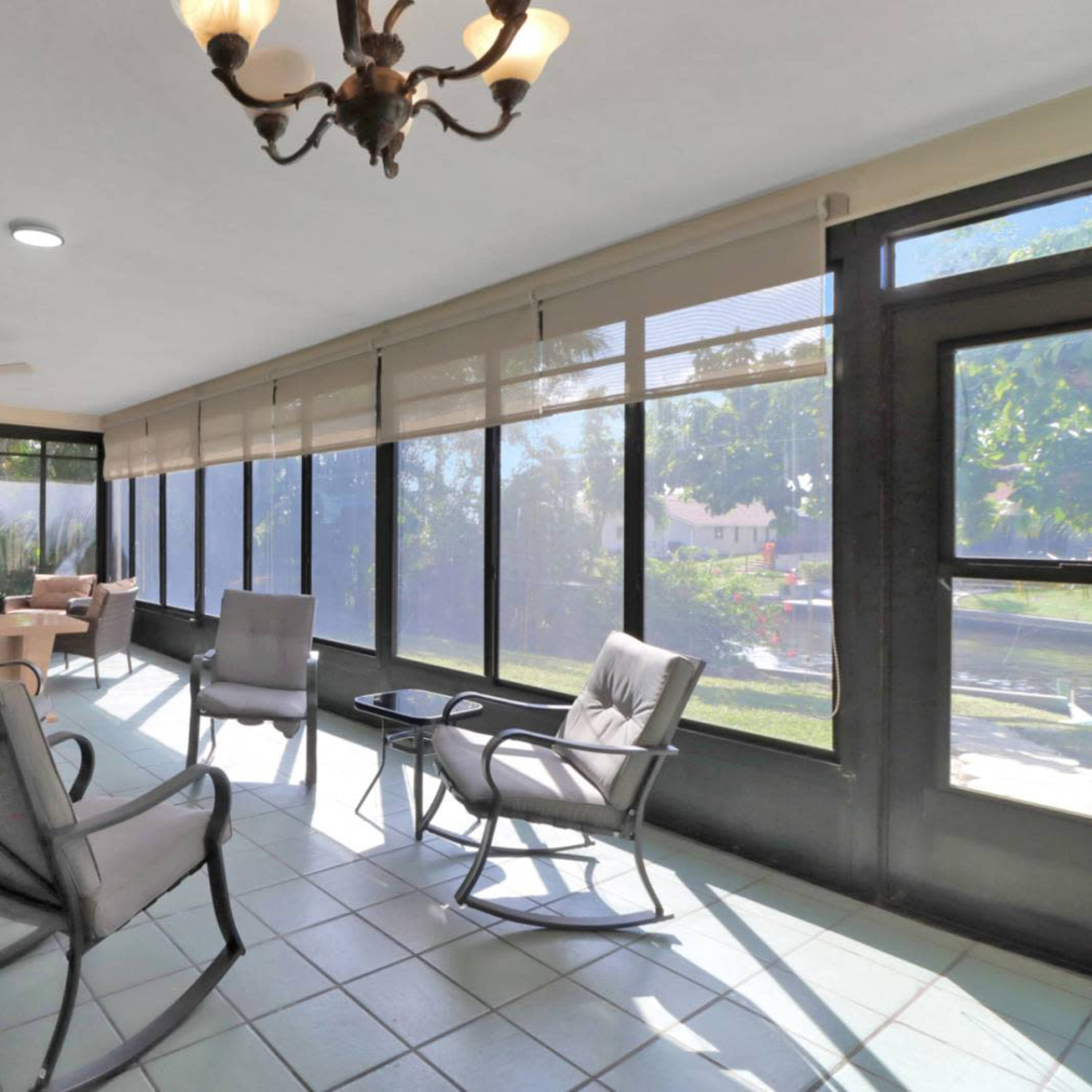 A sunlit screened porch with several chairs, a small table, and large windows displaying a view of greenery outside.