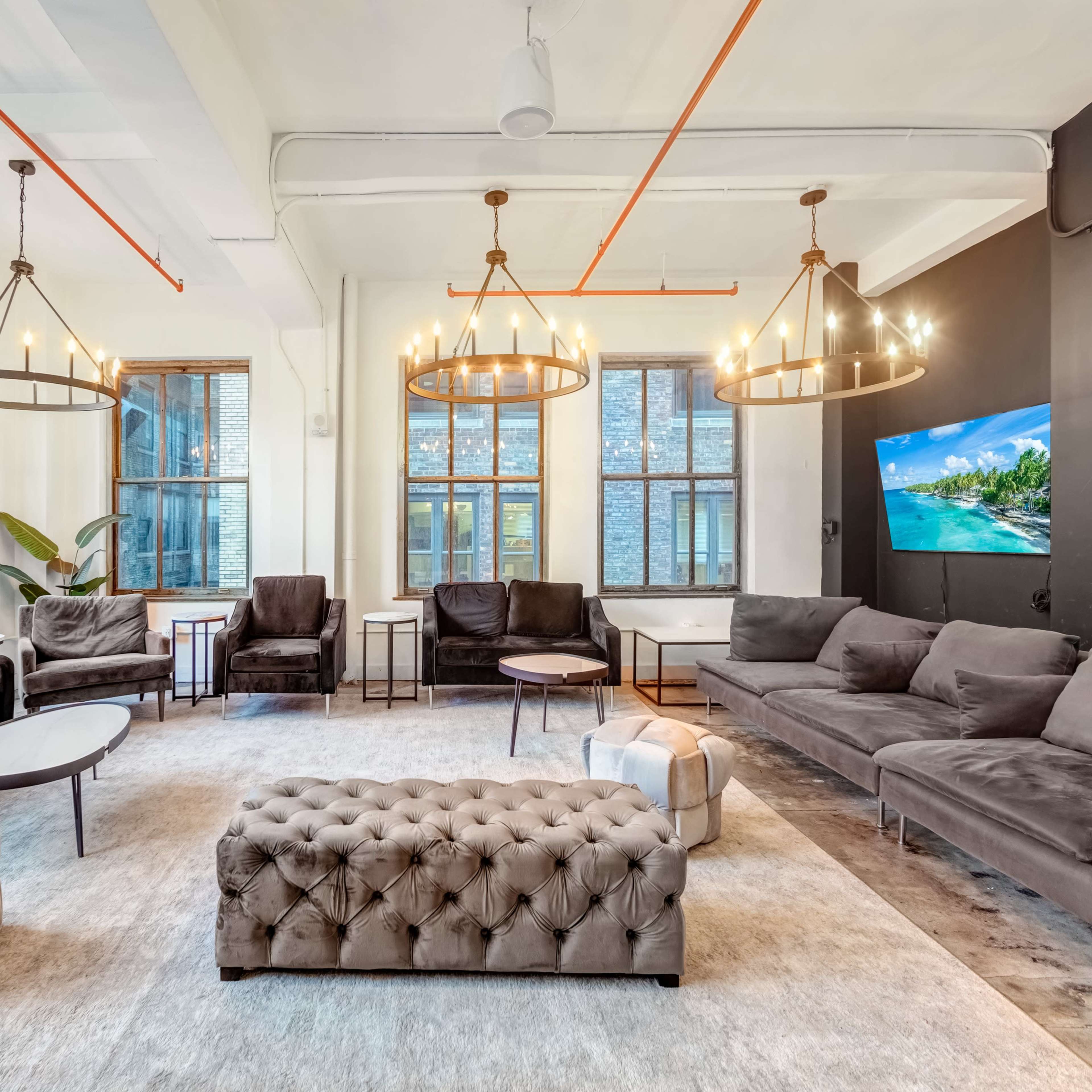 A modern lounge area with large windows, several seating arrangements, a tufted ottoman, and decorative lighting fixtures.