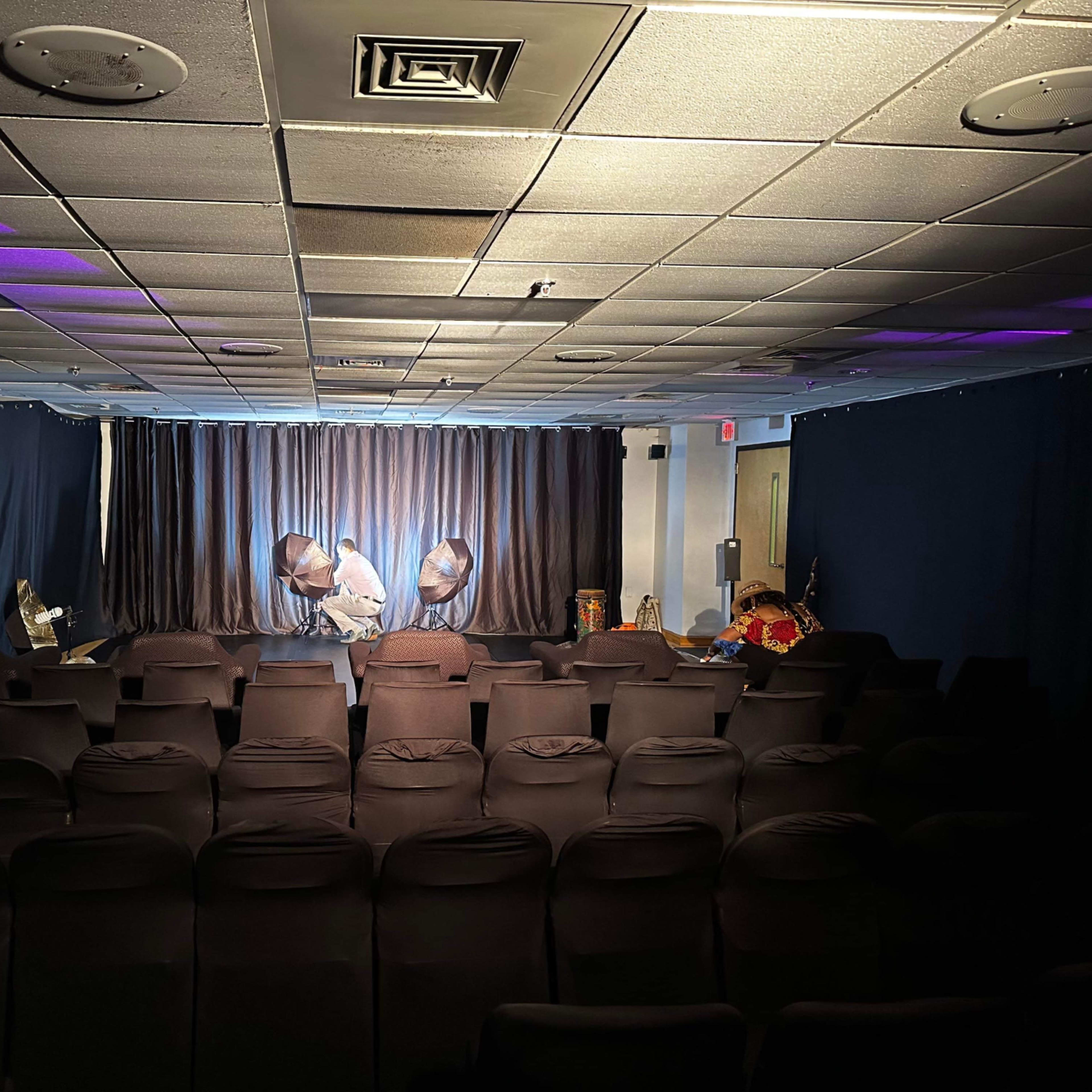 A small auditorium with black chairs facing a stage, which is illuminated by three spotlights and features various props in the background.