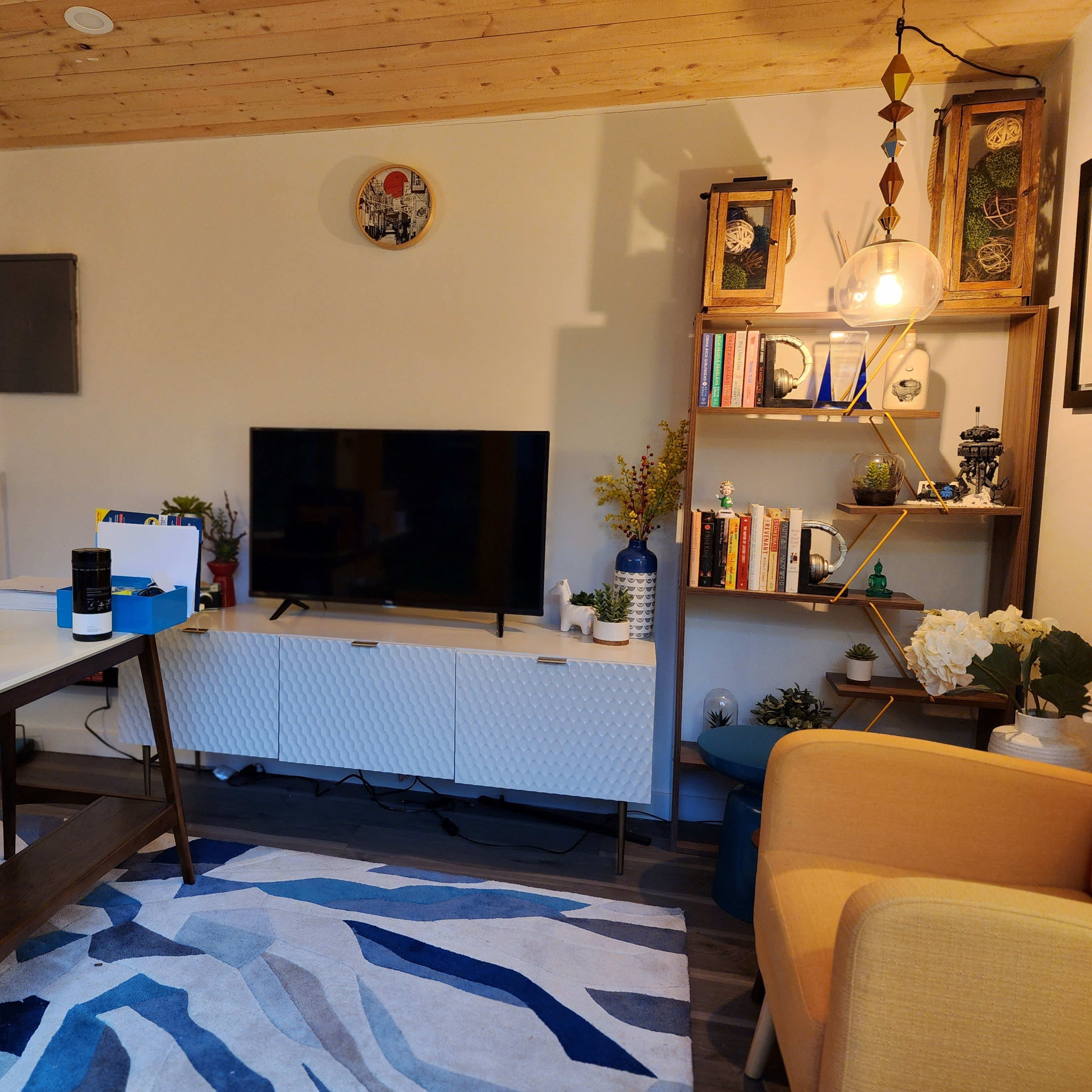 A contemporary living room with a television on a white media console, a wooden desk, an orange armchair, and various decorative items on shelves.