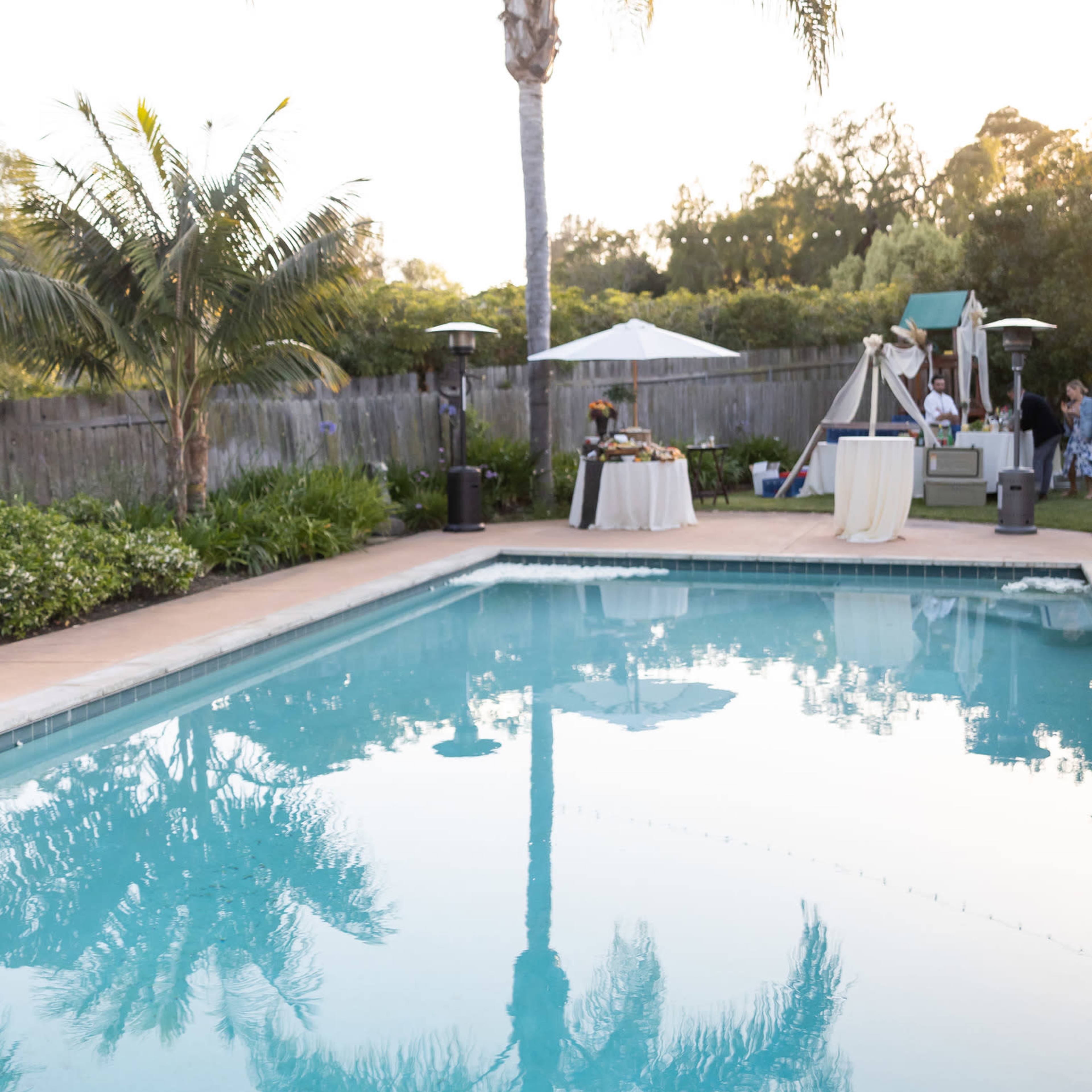 A serene outdoor pool area with palm trees, reflecting the sky and surroundings, accompanied by event setups and gathering spaces nearby.