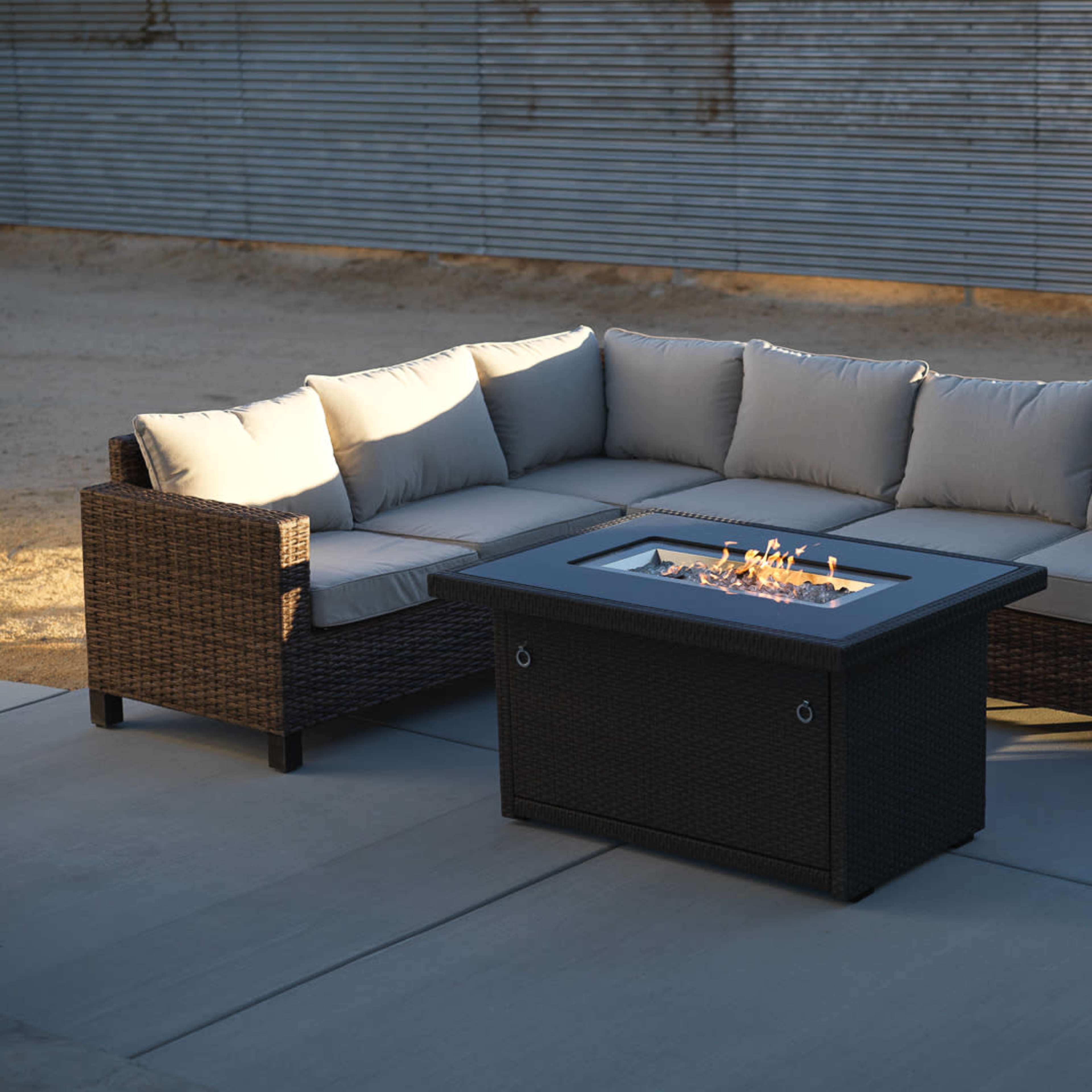 A sectional outdoor couch with beige cushions surrounds a rectangular fire pit on a concrete surface.