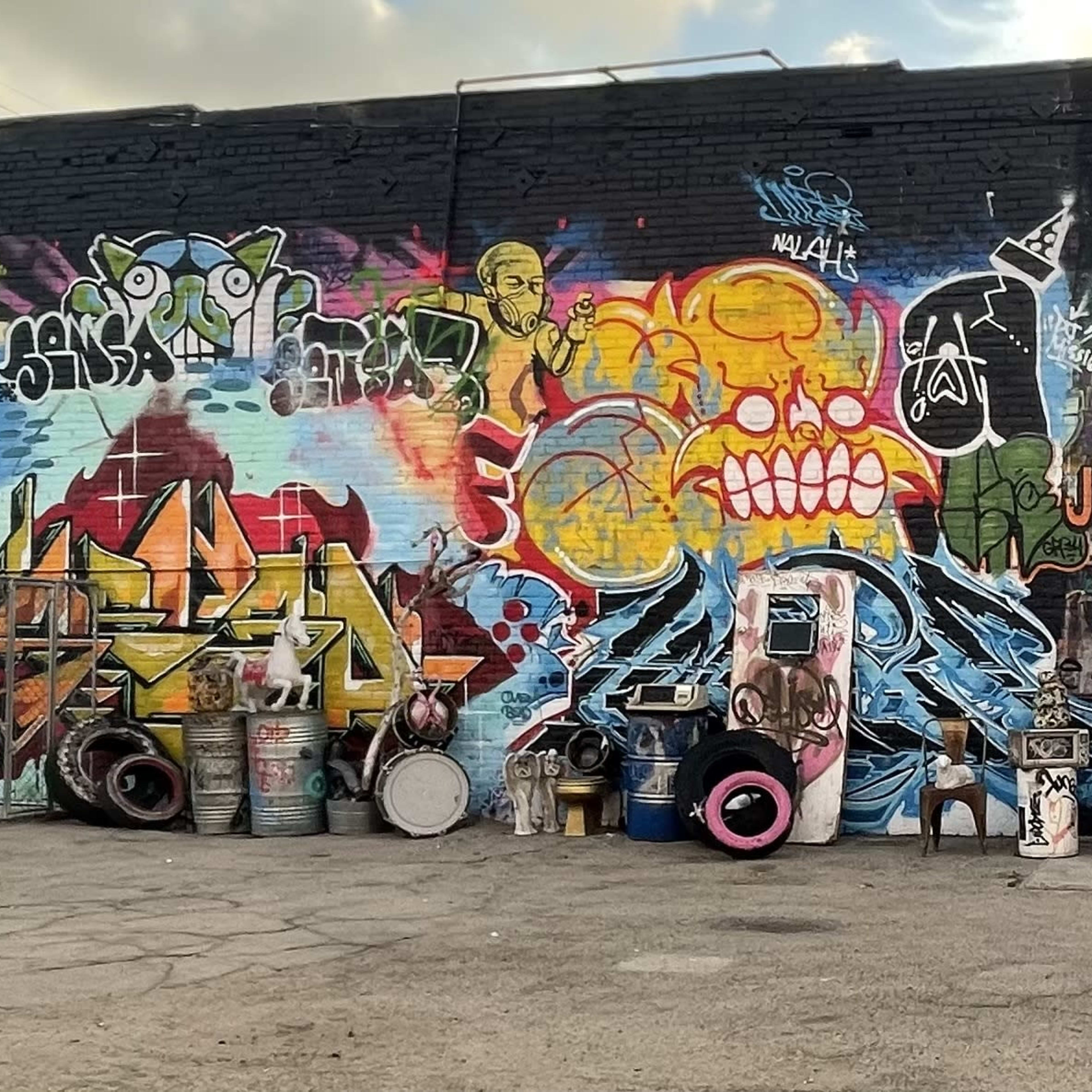 A long wall covered in colorful graffiti art is accompanied by various discarded objects, including tires and old appliances, arranged in front of it.