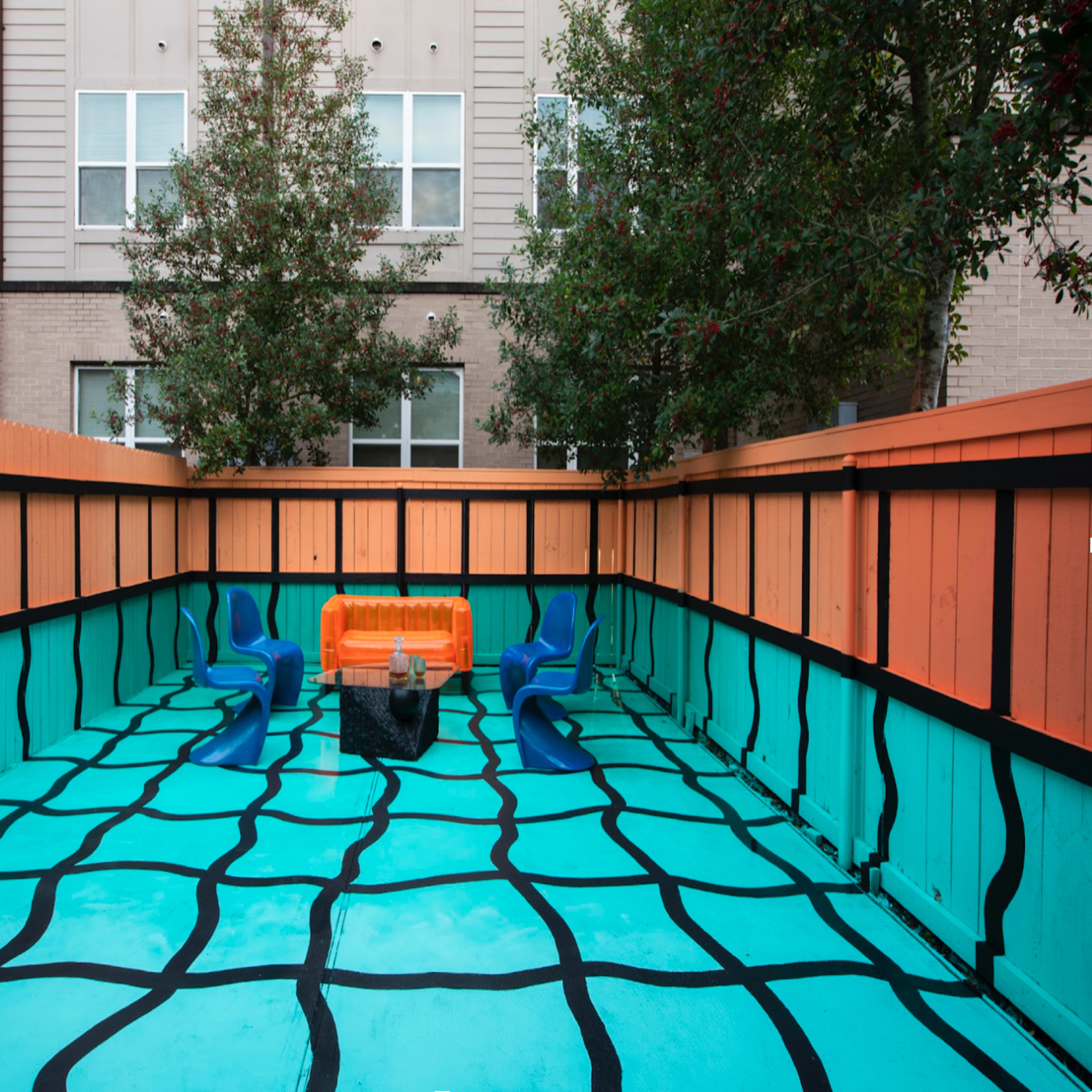 A brightly painted outdoor space with a turquoise floor featuring a black grid pattern, orange benches, and blue chairs surrounded by wooden fencing.