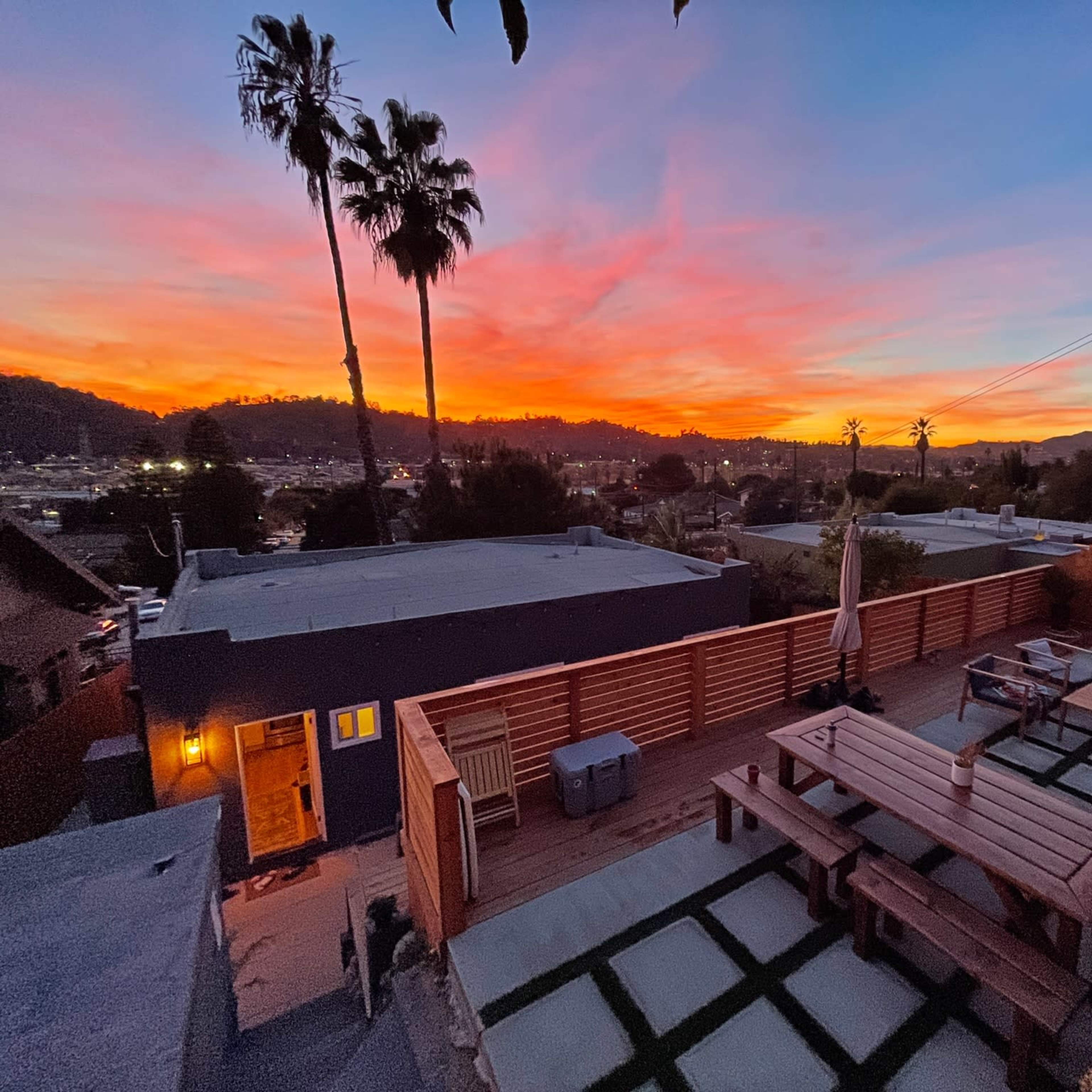 A wooden deck with picnic tables and chairs overlooks a vibrant sunset, with palm trees silhouette against the colorful sky.
