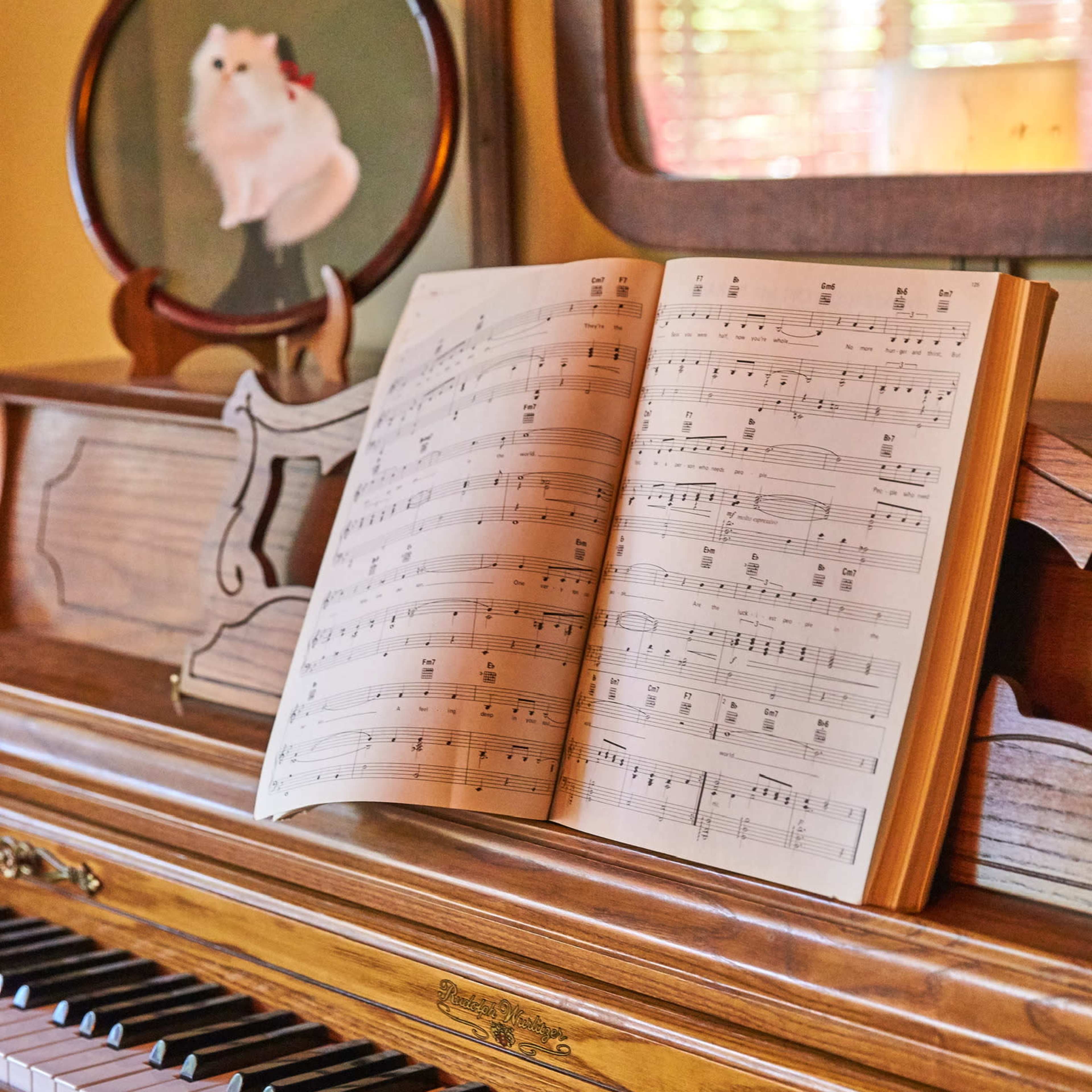 A sheet of music is open on an antique piano, with a circular mirror featuring a small white cat hanging on the wall behind it.
