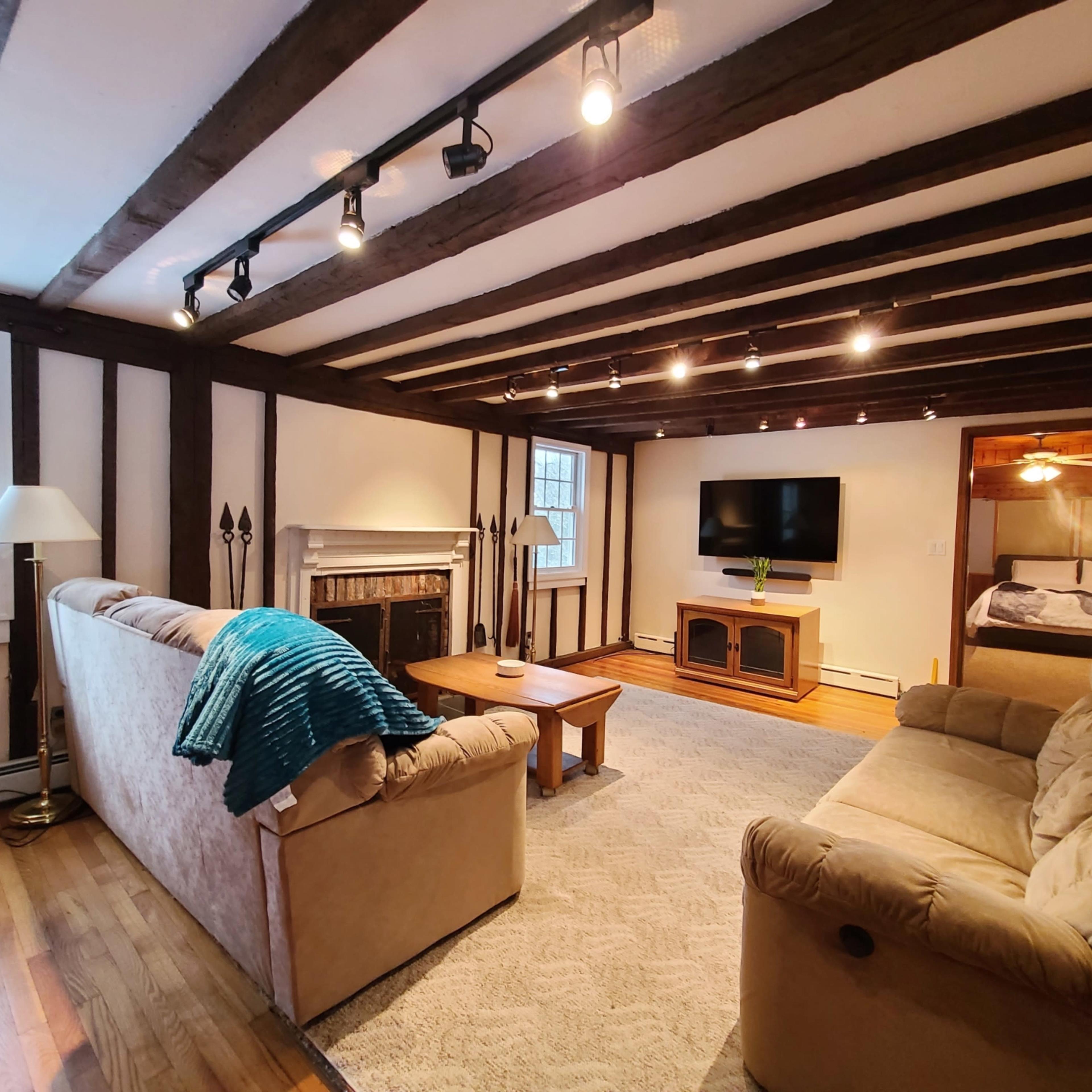 A living room with wooden beams, a fireplace, a flat-screen TV mounted on the wall, and a doorway leading to a bedroom.
