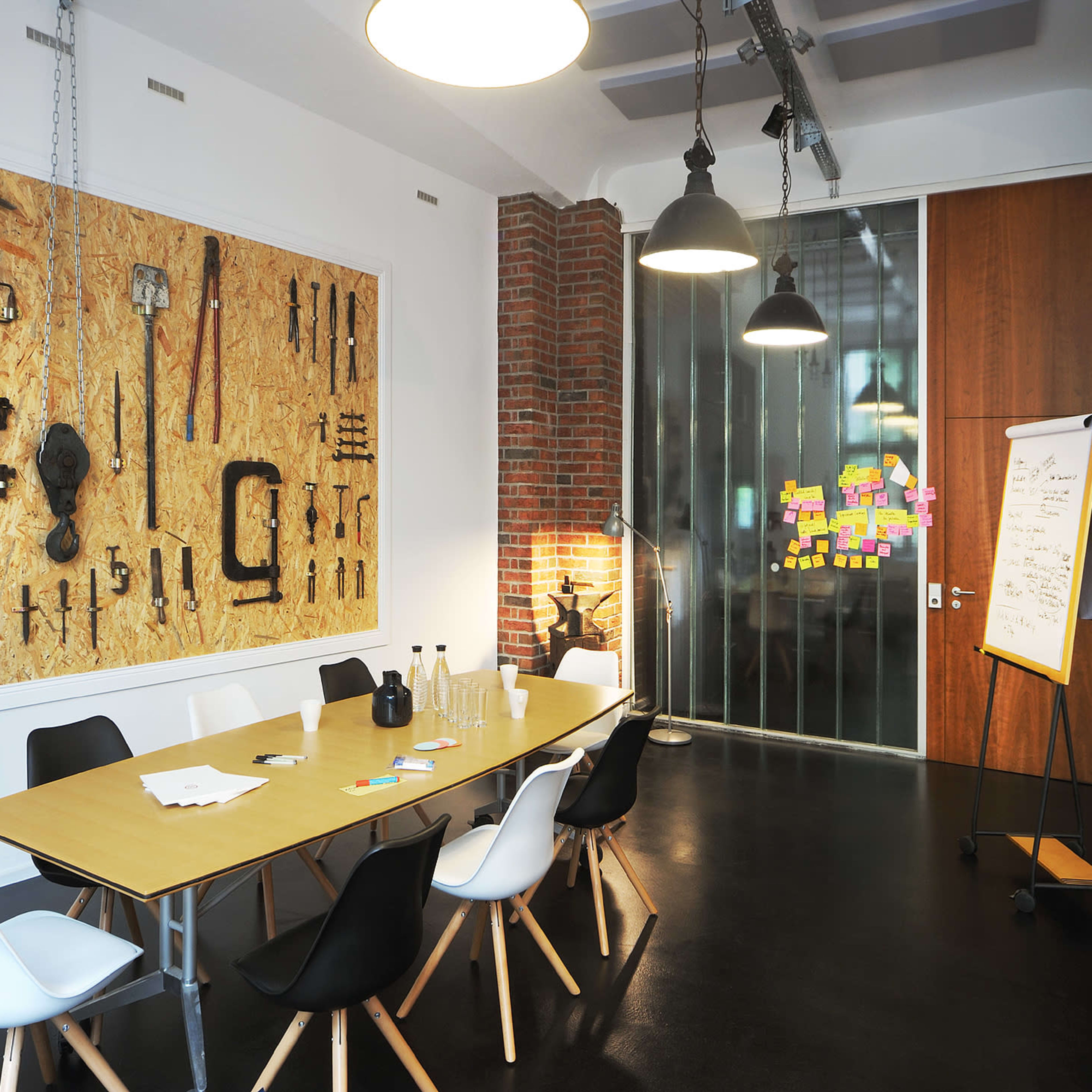 A modern meeting room features a central wooden table surrounded by black and white chairs, with a wall adorned by various tools and a glass partition displaying colorful notes.