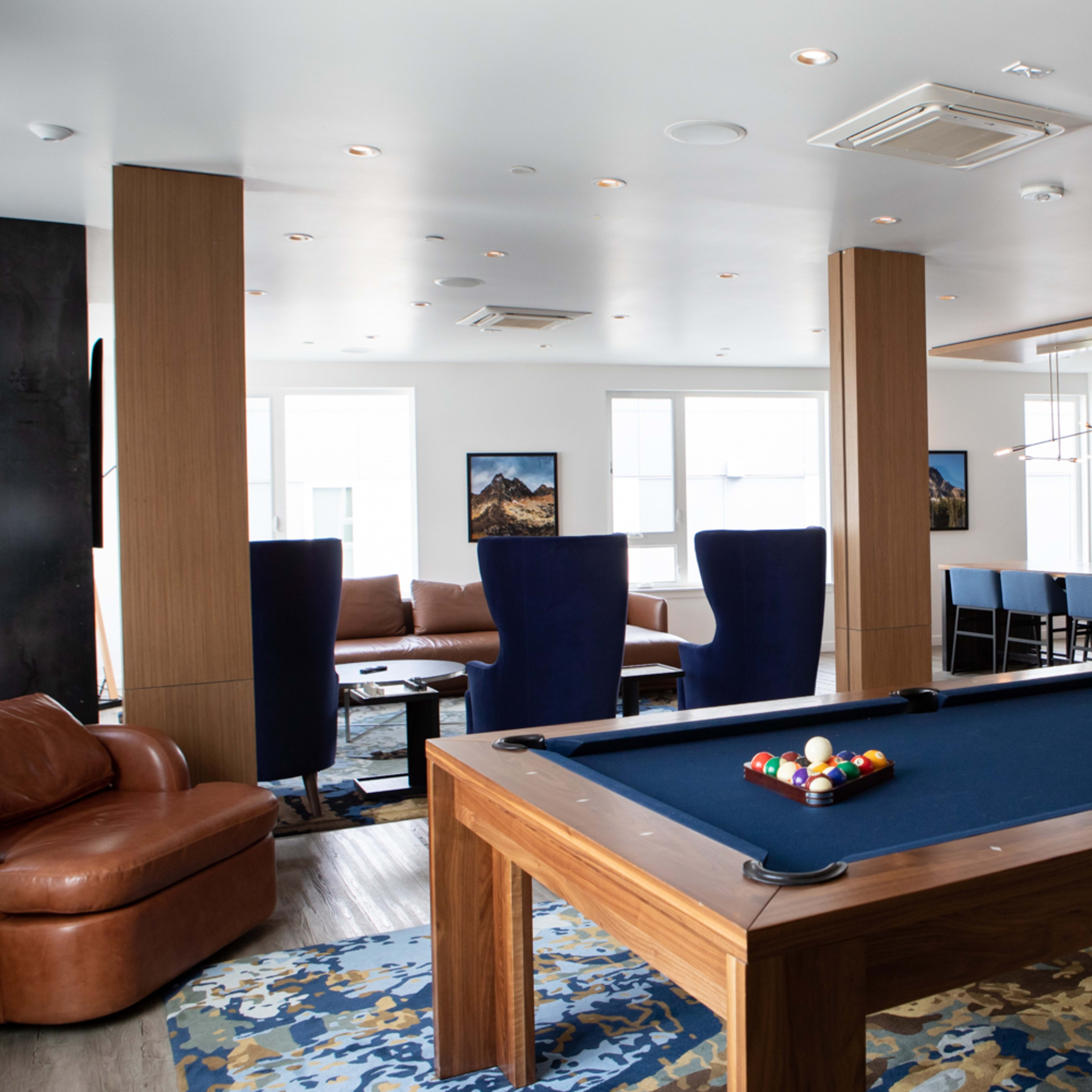 A modern game room featuring a billiards table, a seating area with leather sofas, and a kitchen in the background.