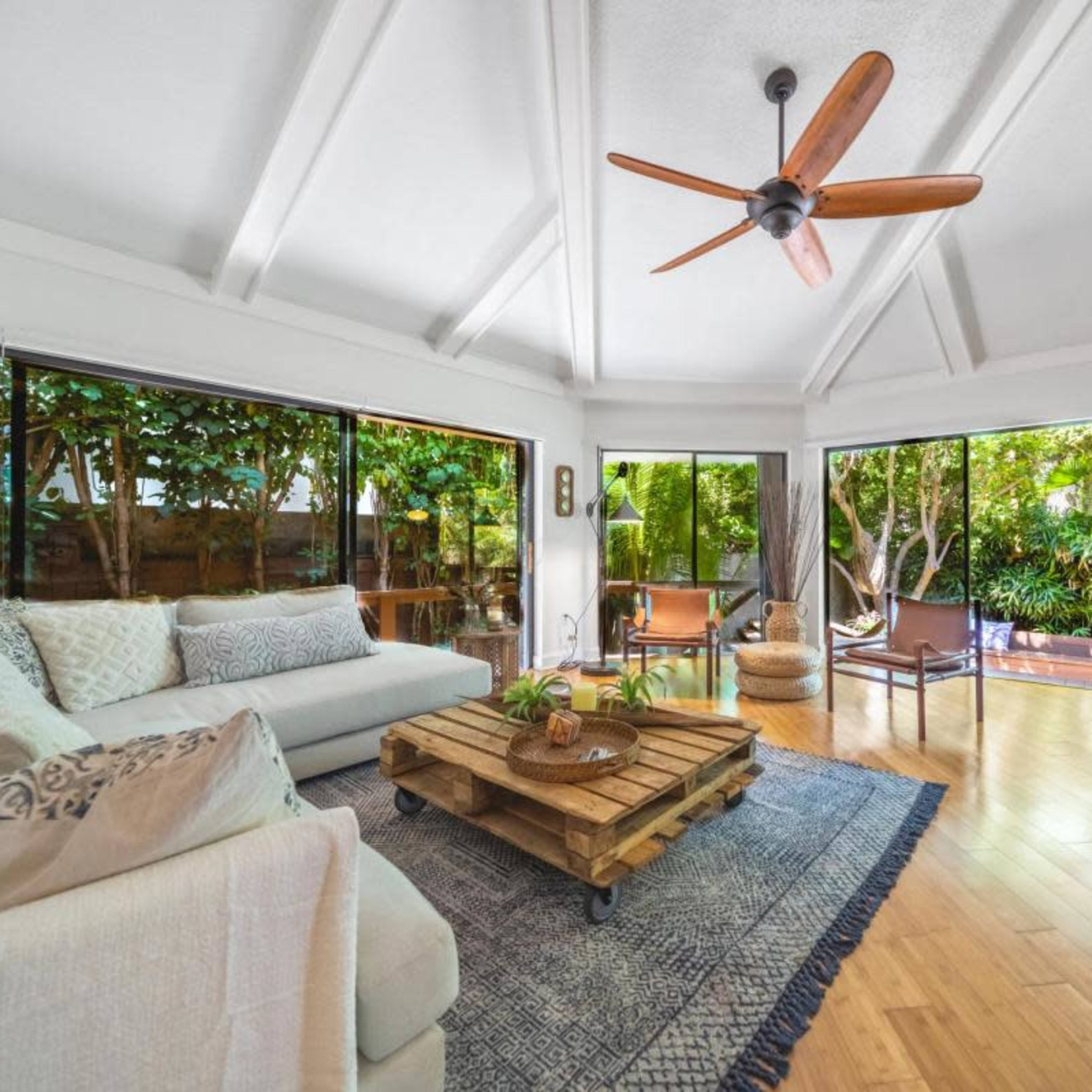 A bright living room with a wooden coffee table, a cozy sofa, and large glass windows overlooking lush greenery.