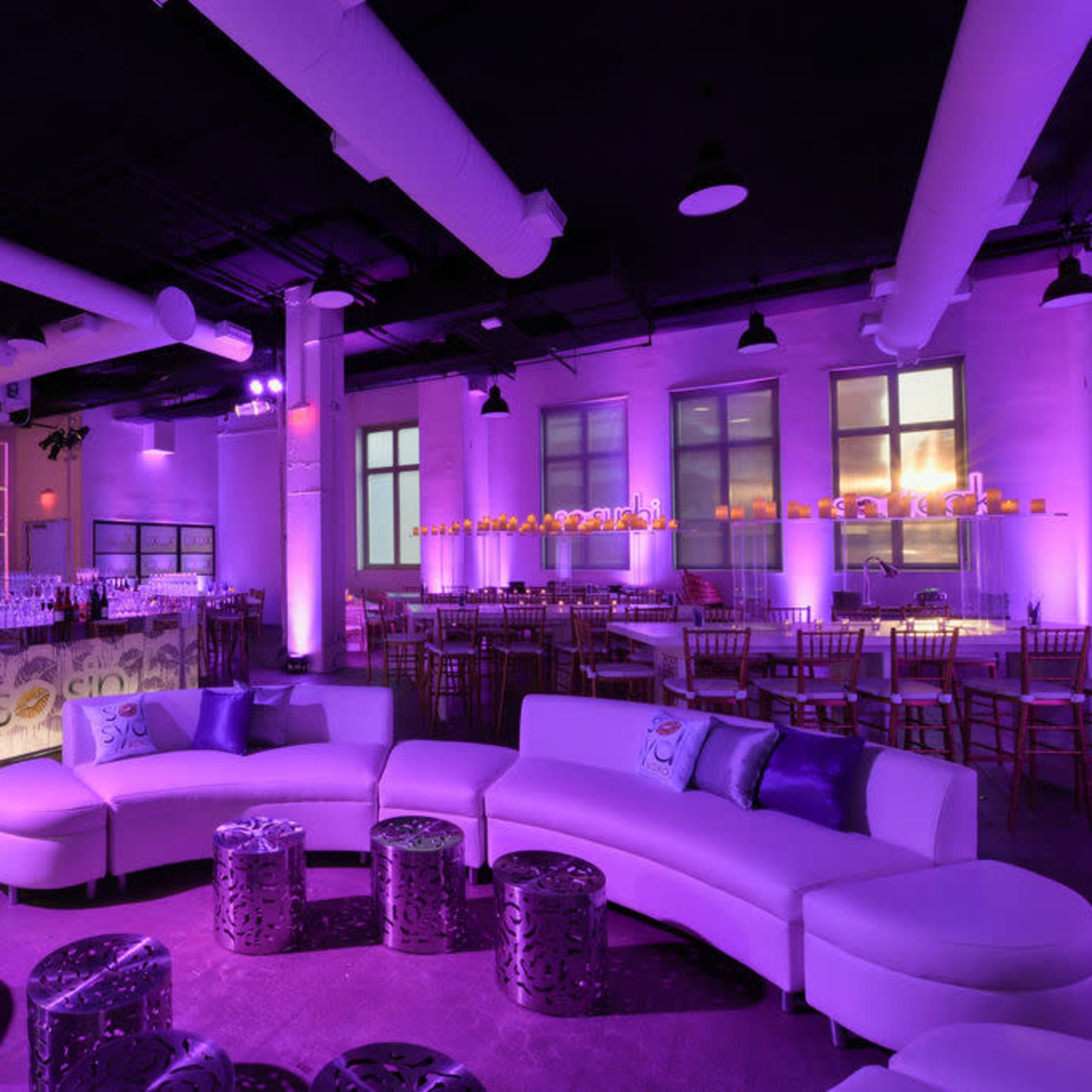 A modern event space with purple lighting, featuring a curved white sofa, tables, and a backlit bar.