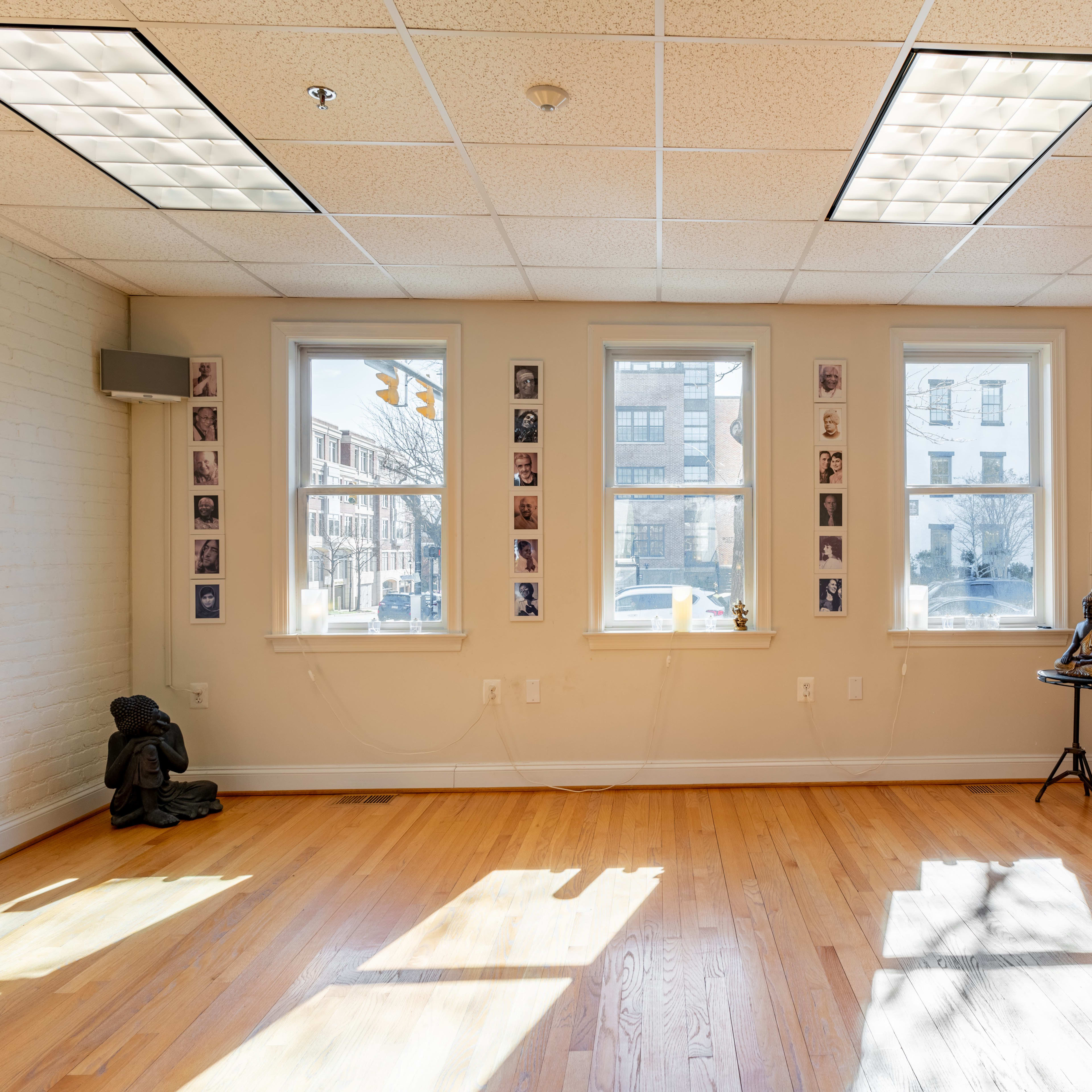 A well-lit room with wooden floors, three windows flanked by photographs on the walls, and a small sculpture in the corner.