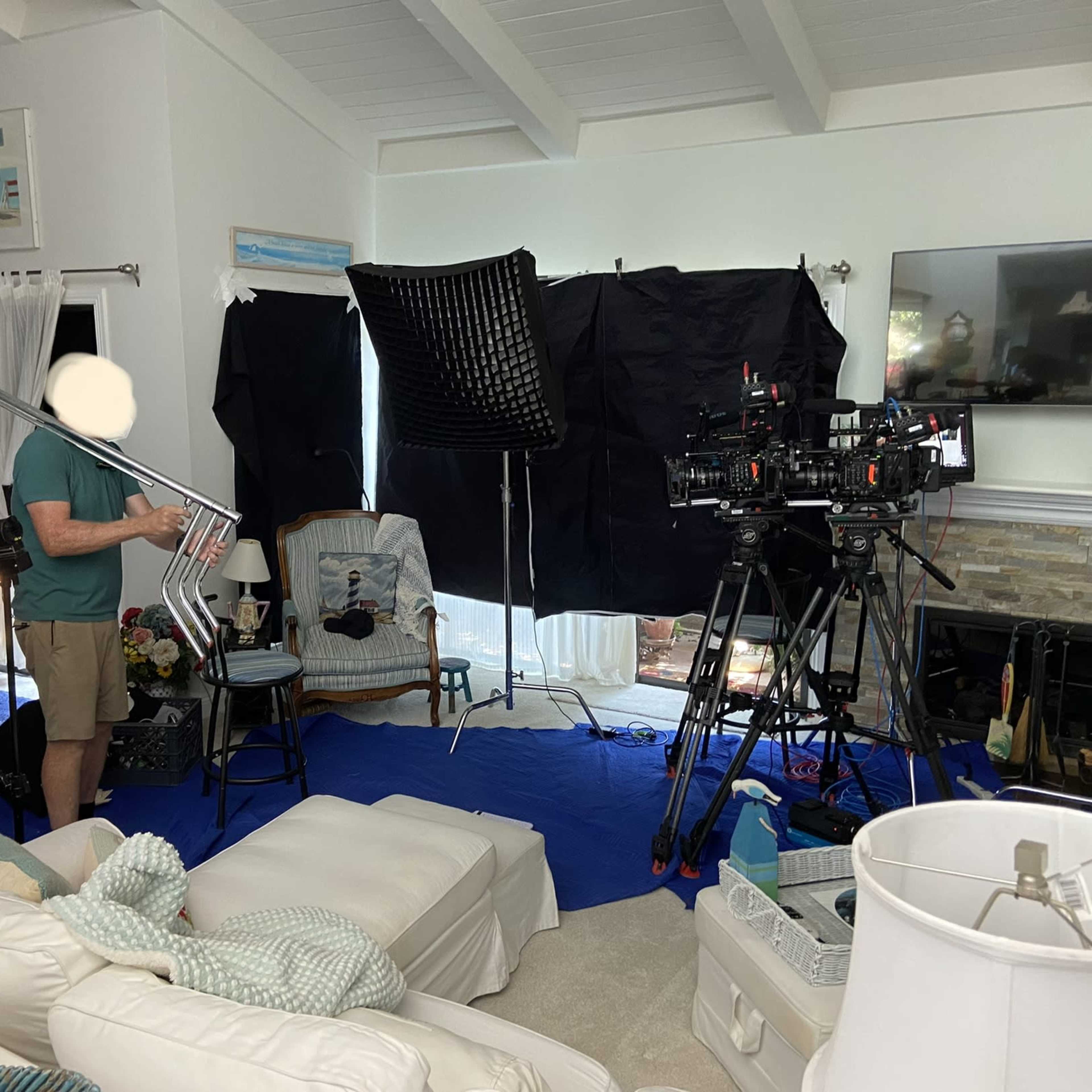 A film crew setting up equipment in a brightly lit living room, with cameras, lights, and sound equipment arranged around a seating area.