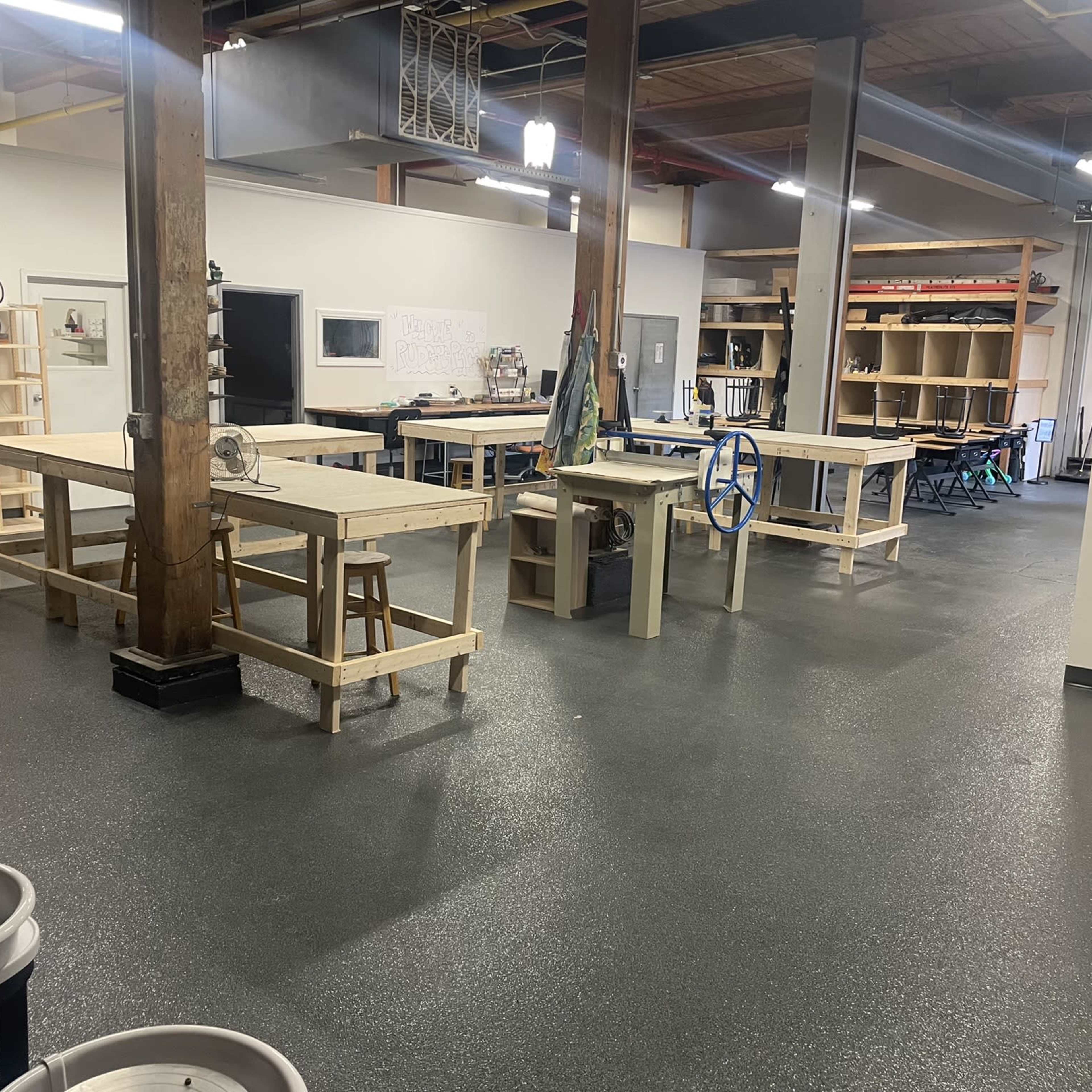 An indoor workspace with multiple wooden tables, shelves, and pottery wheels, all arranged neatly in a well-lit room.