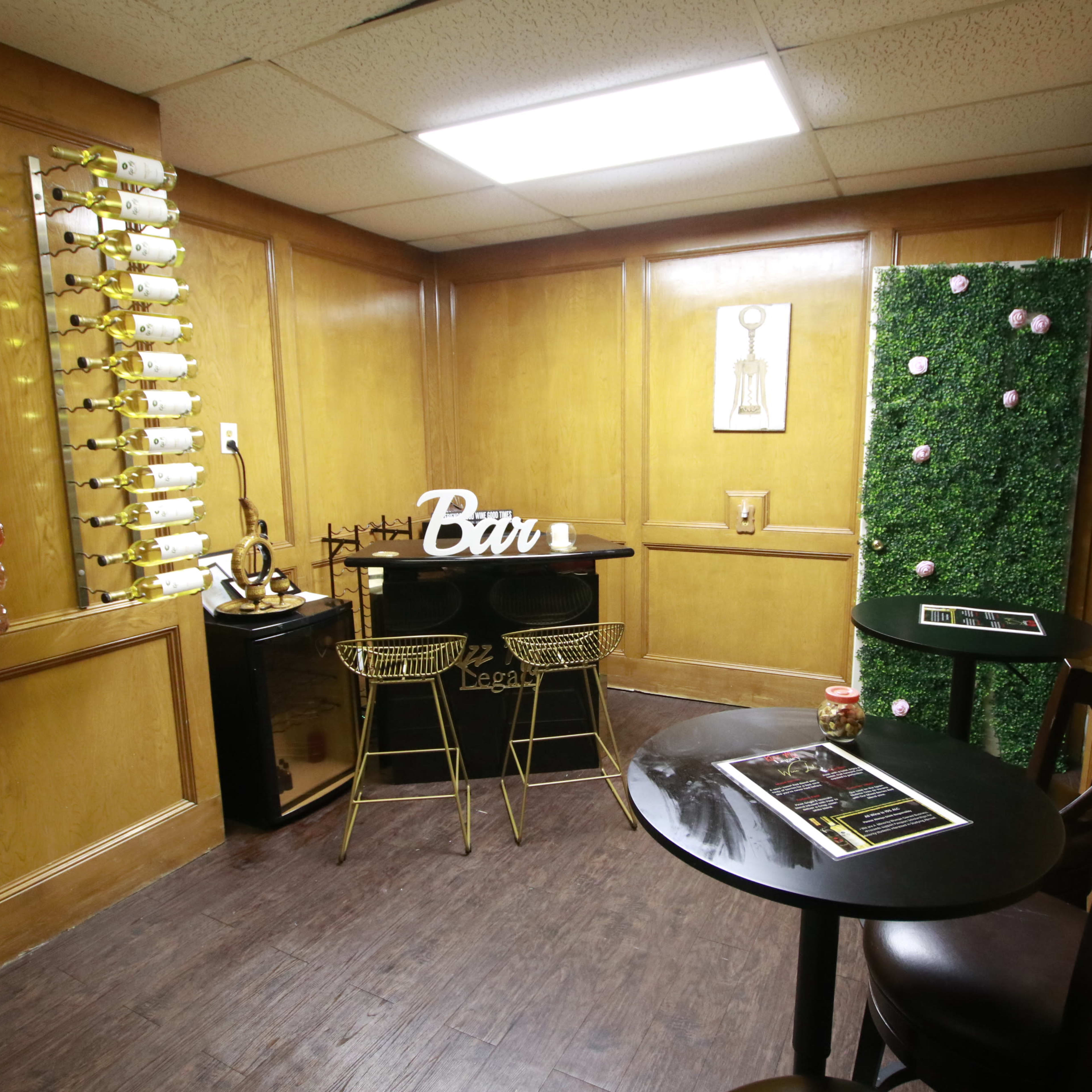 A small wooden-paneled room featuring wine racks on one wall, a black bar table with gold stools, and a decorative green wall.