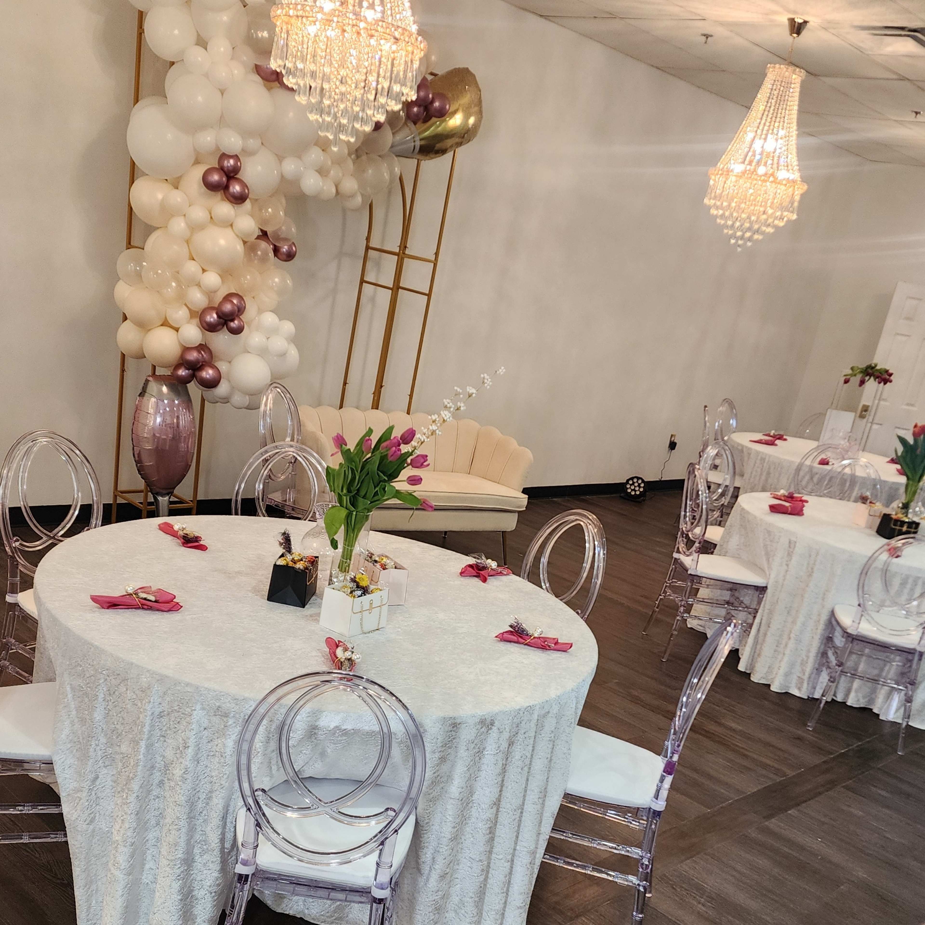 A decorated event space with round tables covered in white tablecloths, clear acrylic chairs, and a backdrop of balloons and chandeliers.