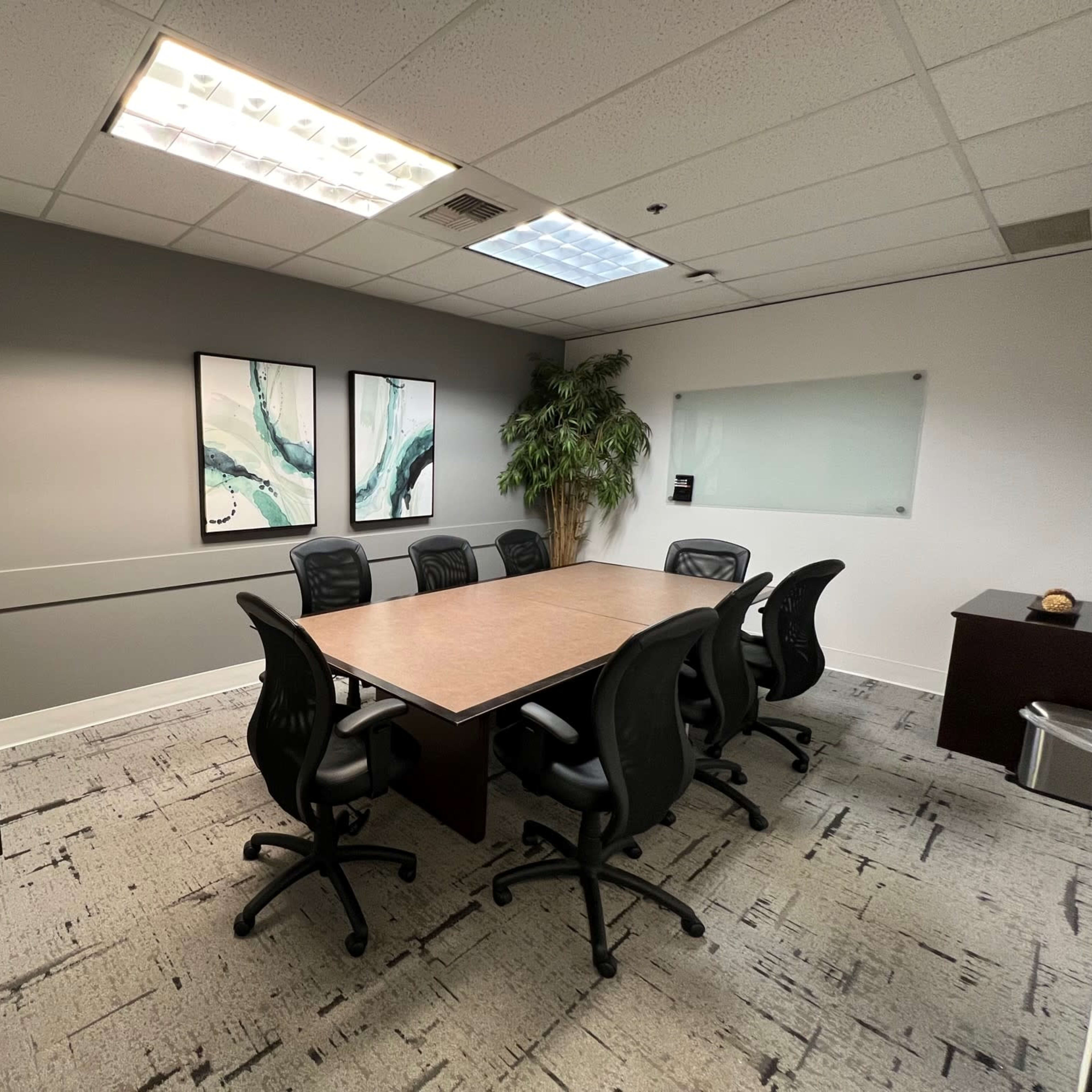 A conference room features a long wooden table surrounded by black office chairs, with two abstract paintings on the wall and a plant in one corner.