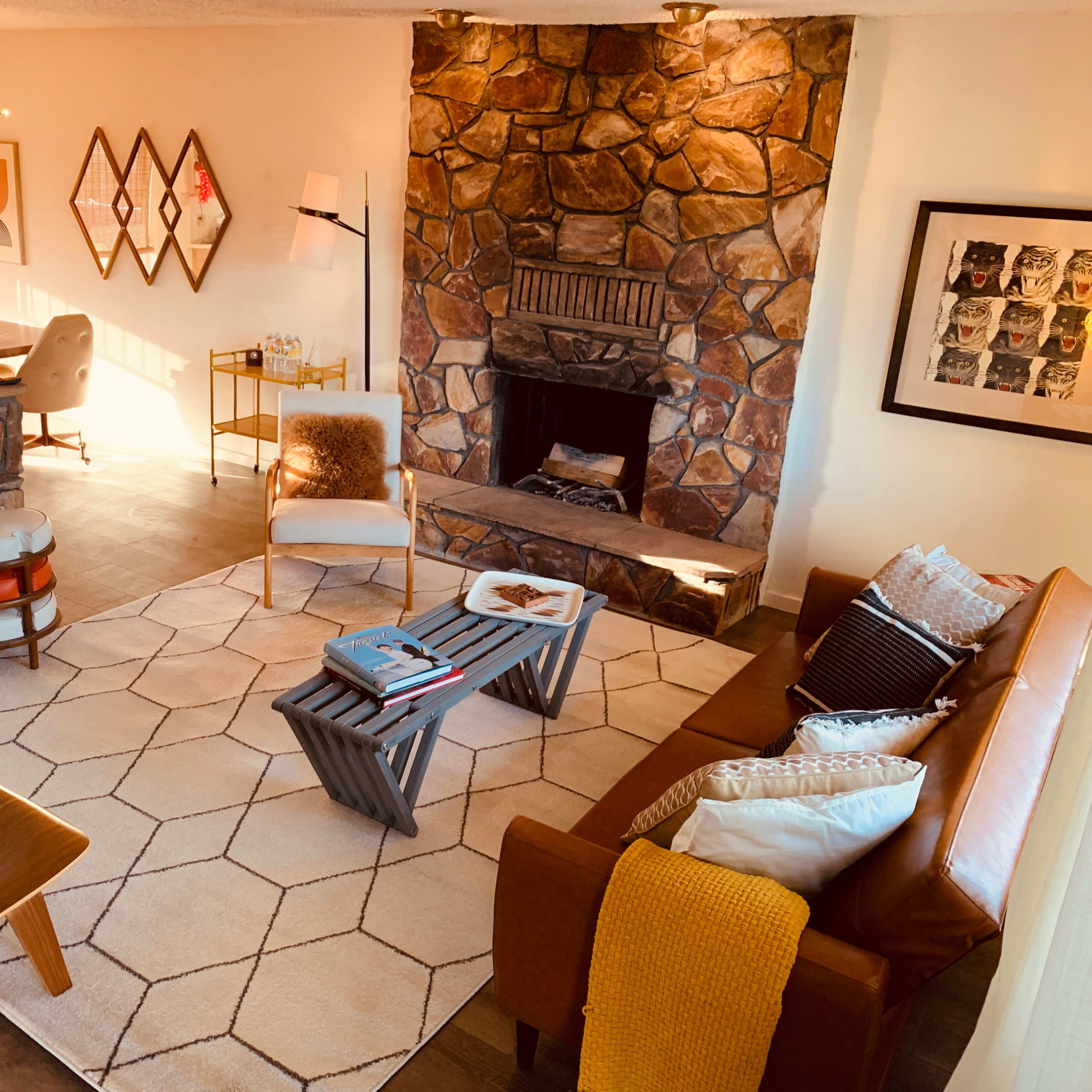 A cozy living room featuring a stone fireplace, a brown leather sofa, a geometric patterned rug, and wooden accent chairs.