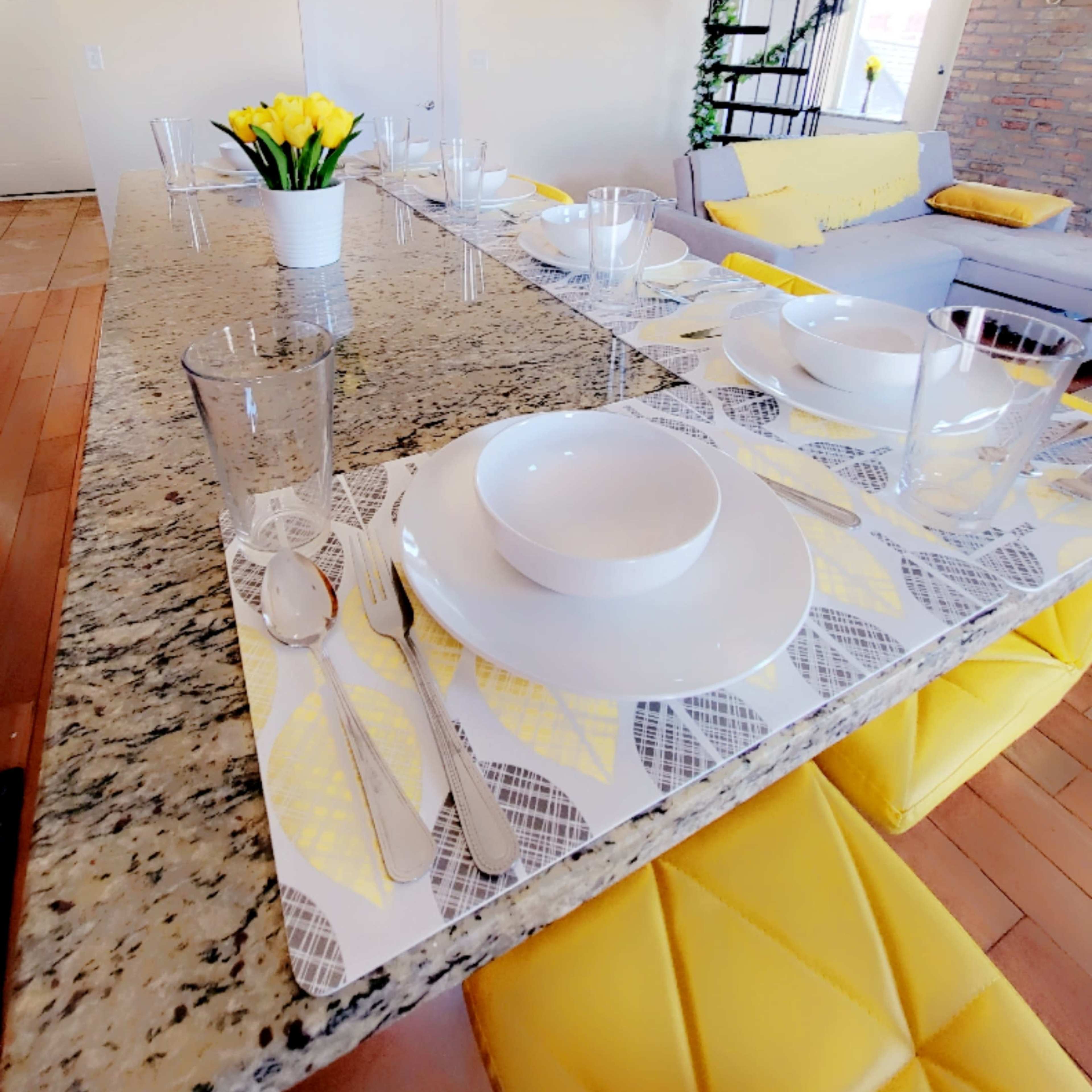 A dining table is set with white plates and glasses on a patterned placemat, alongside a vase of flowers, in a bright room featuring a spiral staircase and a yellow-accented sofa.