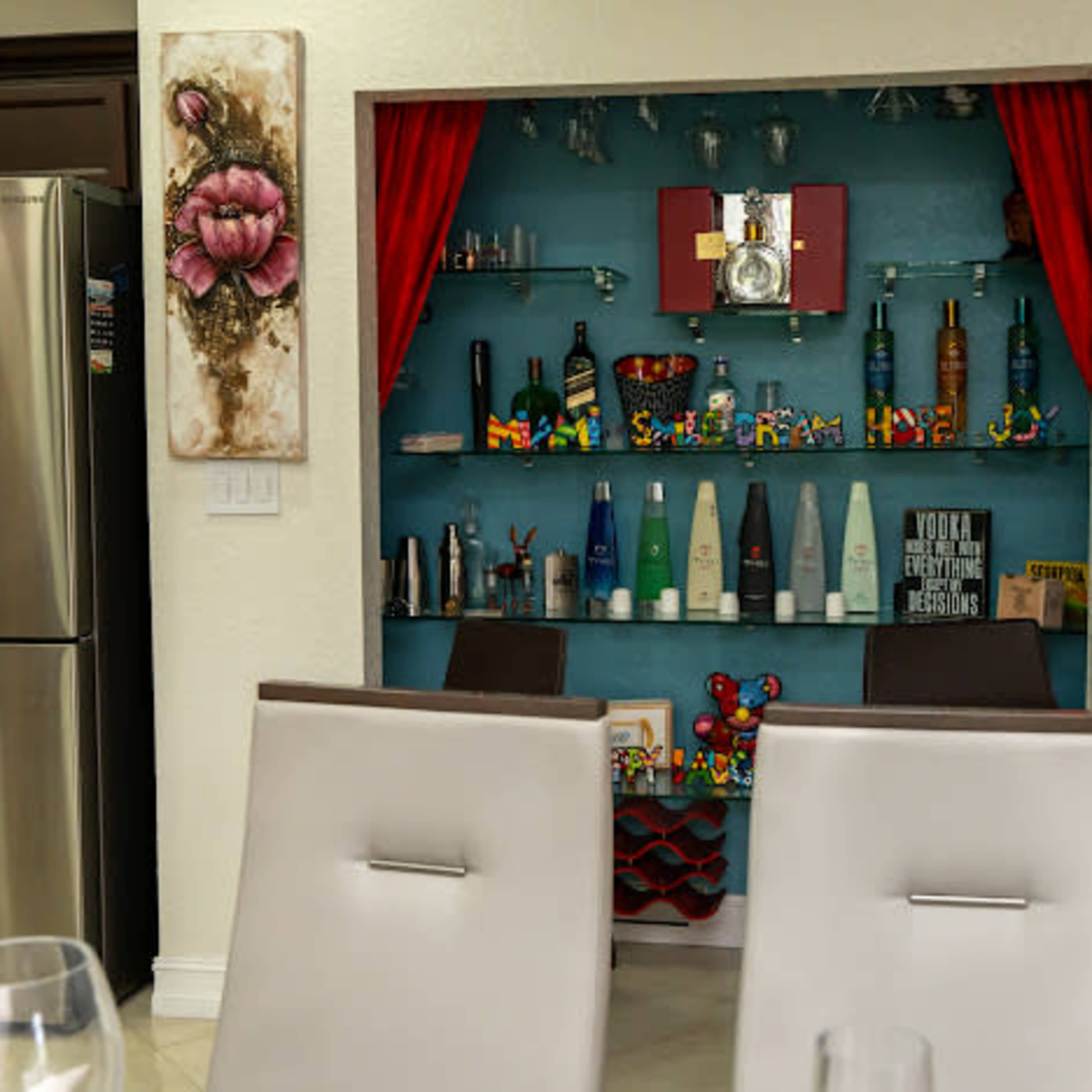 A home bar area with decorative shelves displaying various bottles and colorful toys, framed by red curtains and accompanied by wall art.