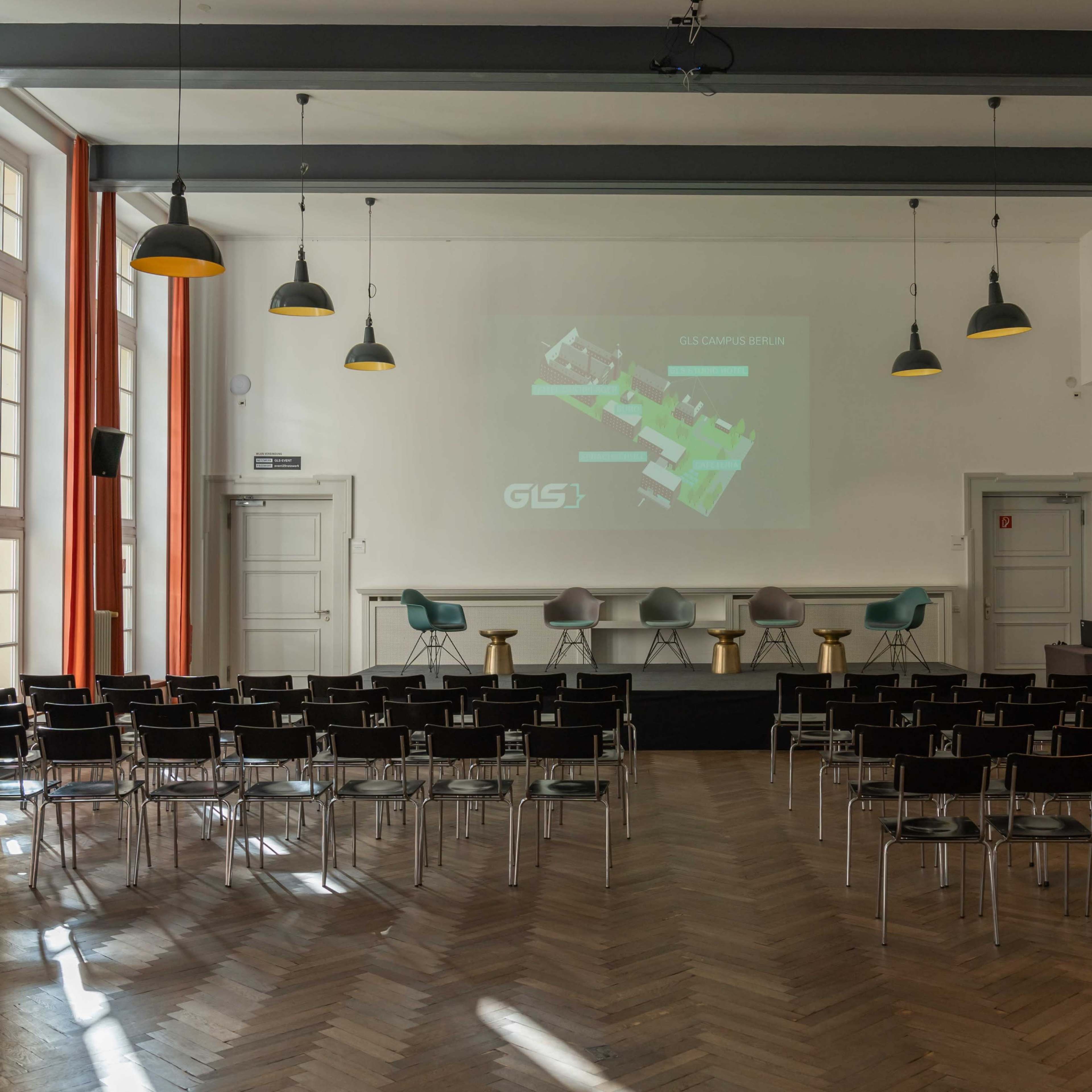A conference room setup with rows of chairs facing a projector screen displaying a map, accompanied by a small stage with stools.