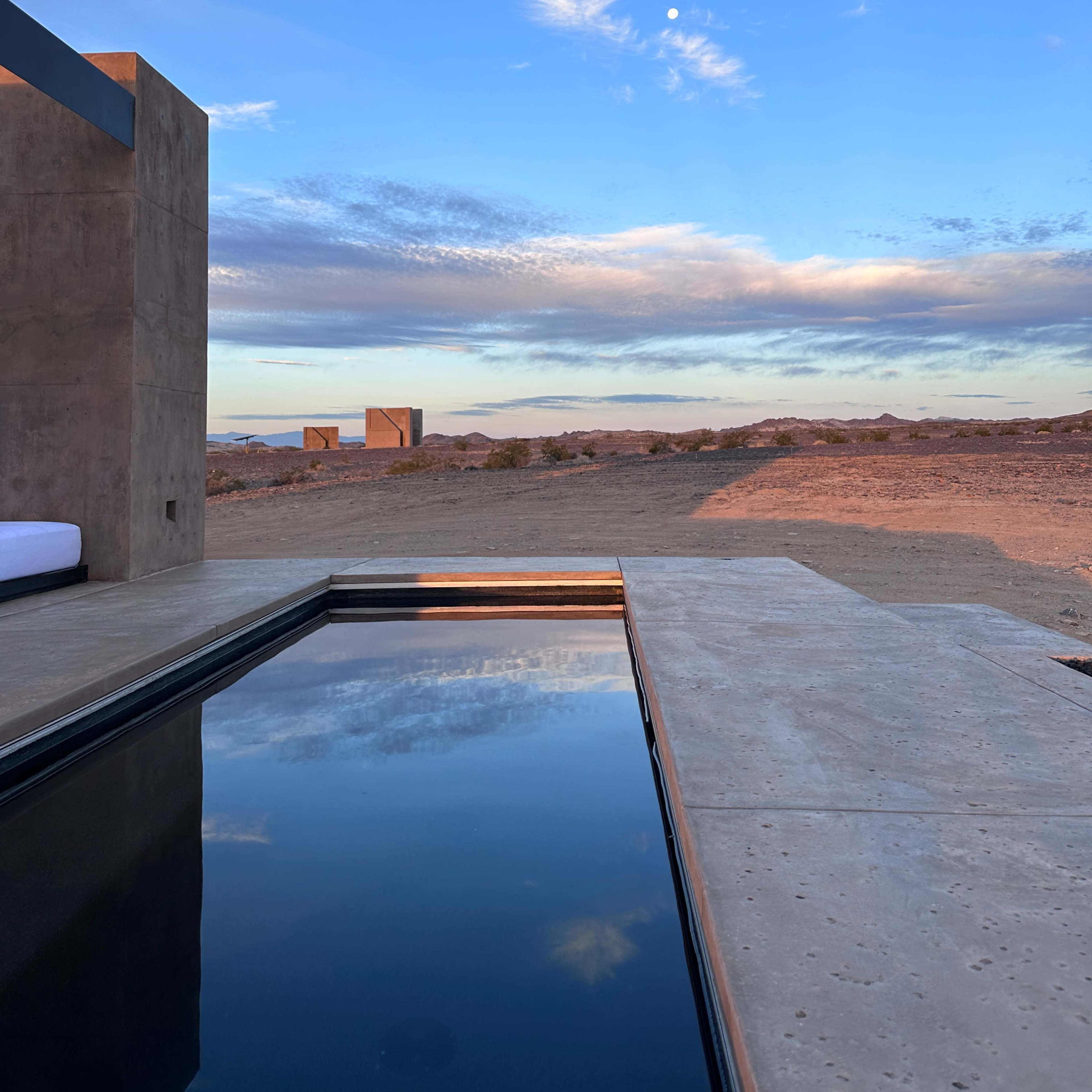 A minimalist pool reflecting the sky and landscape in a desert setting with distant structures and clouds.