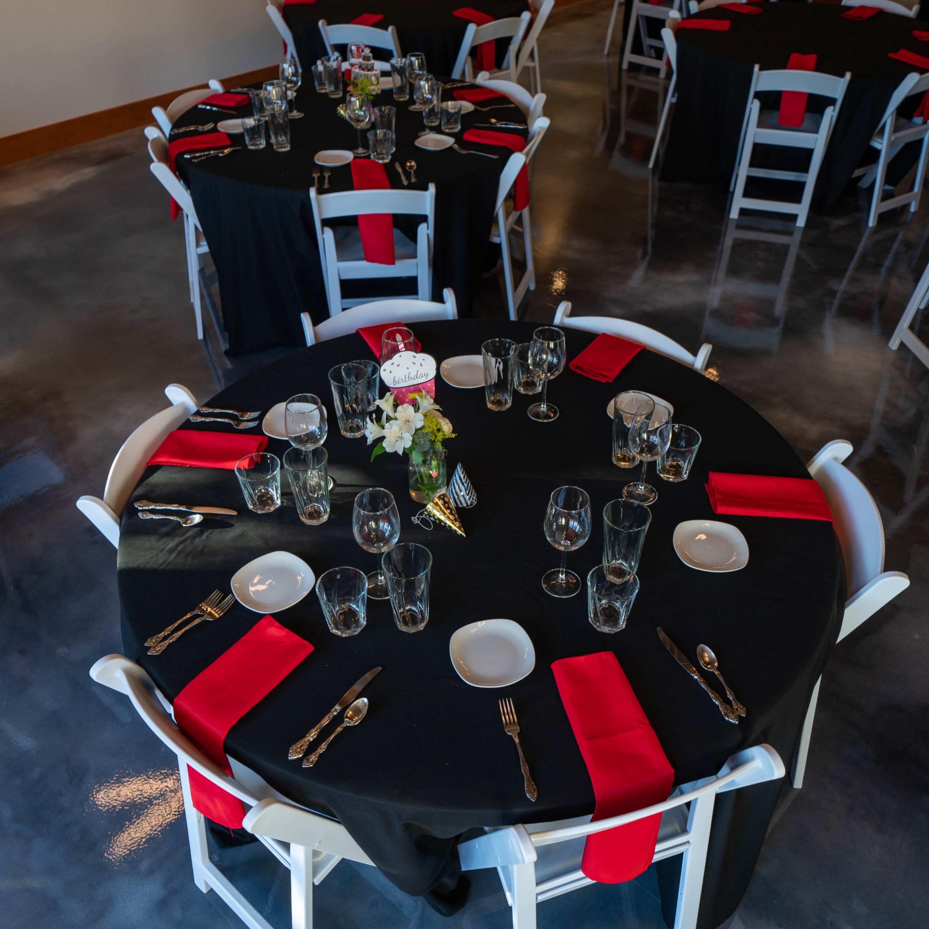A banquet hall is set up with round tables covered in black cloth, featuring red napkins, glassware, and silverware.