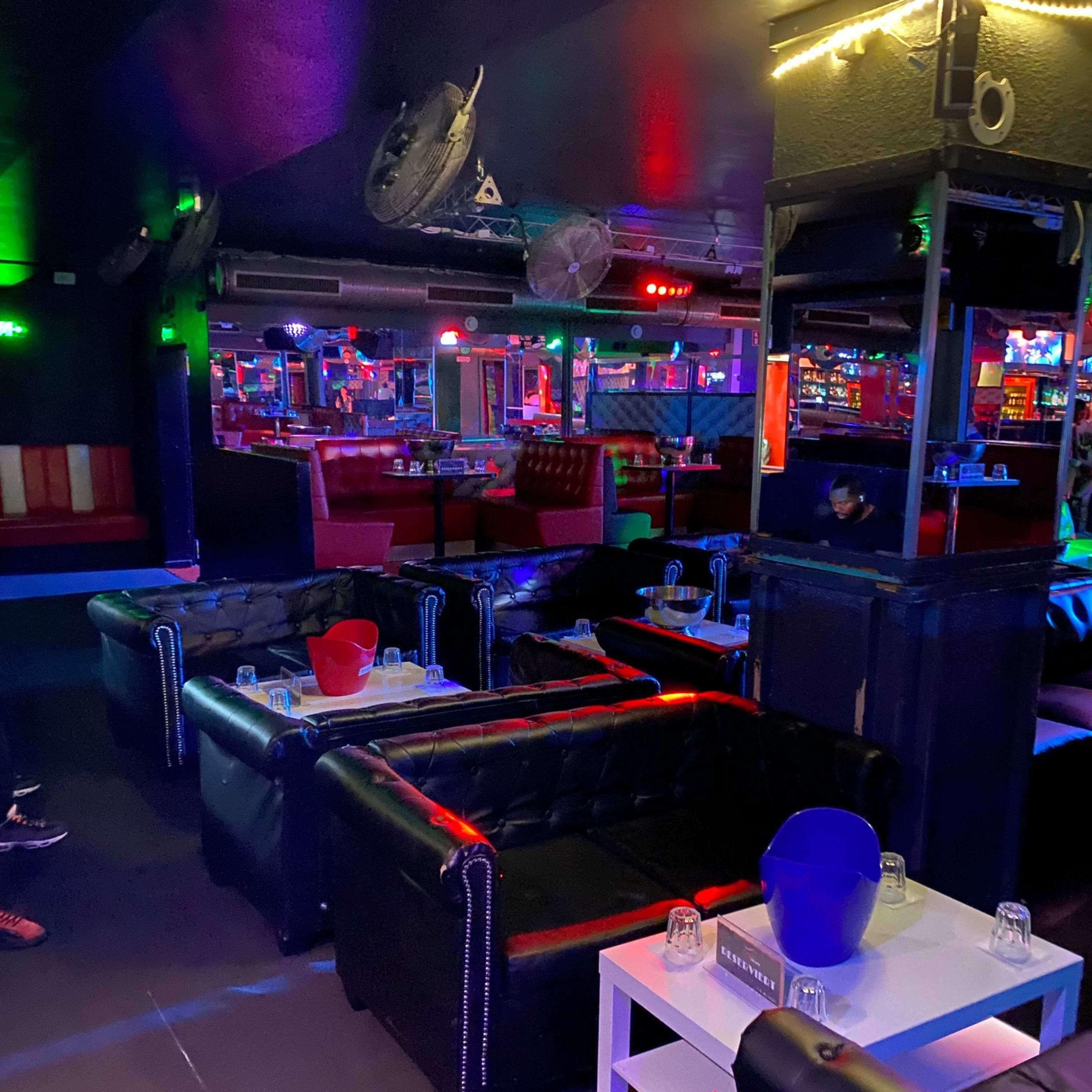 A dimly lit lounge with black leather couches, small tables, and colorful lighting in the background.