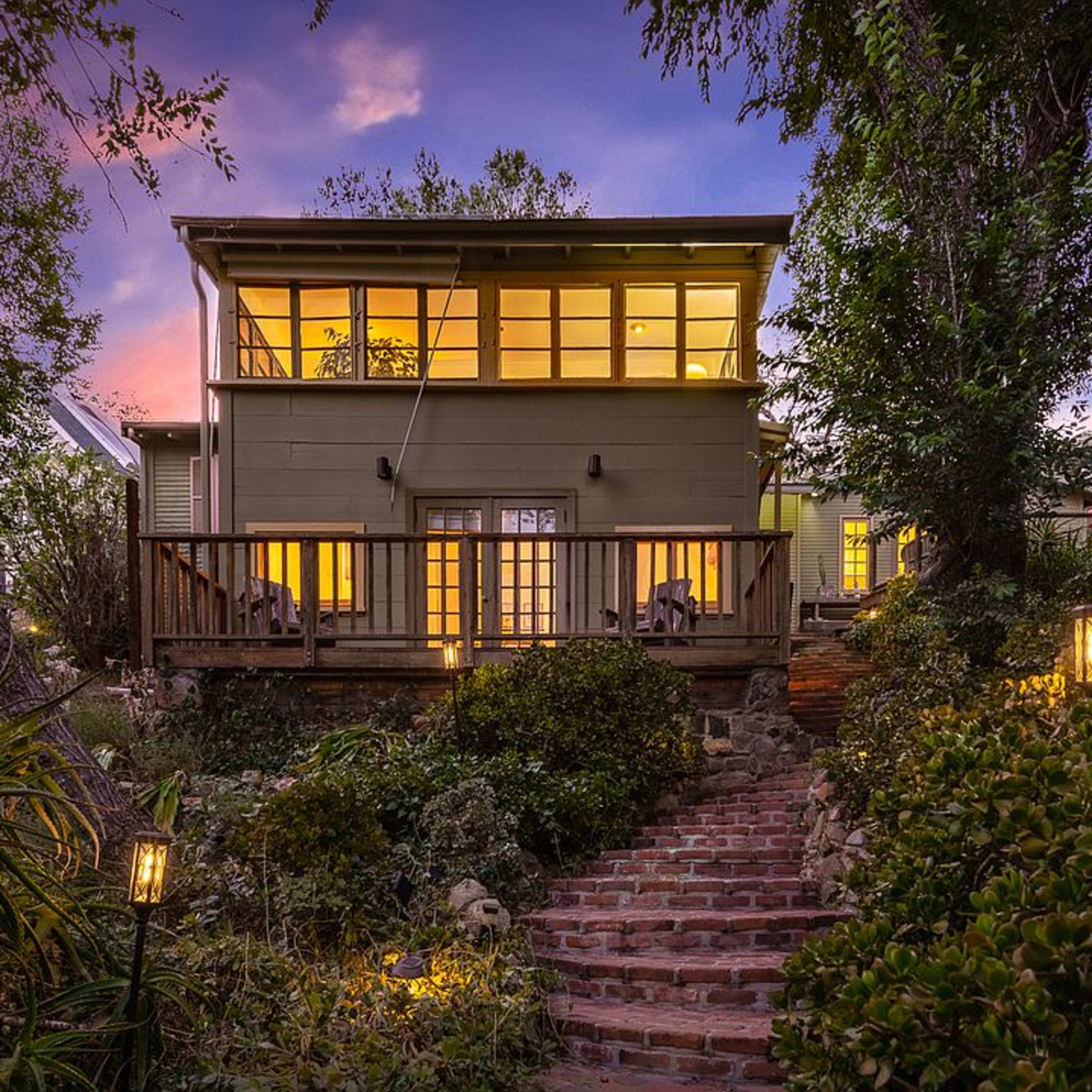A two-story green house is surrounded by garden plants, with a brick pathway leading to a wooden deck illuminated by outdoor lights.