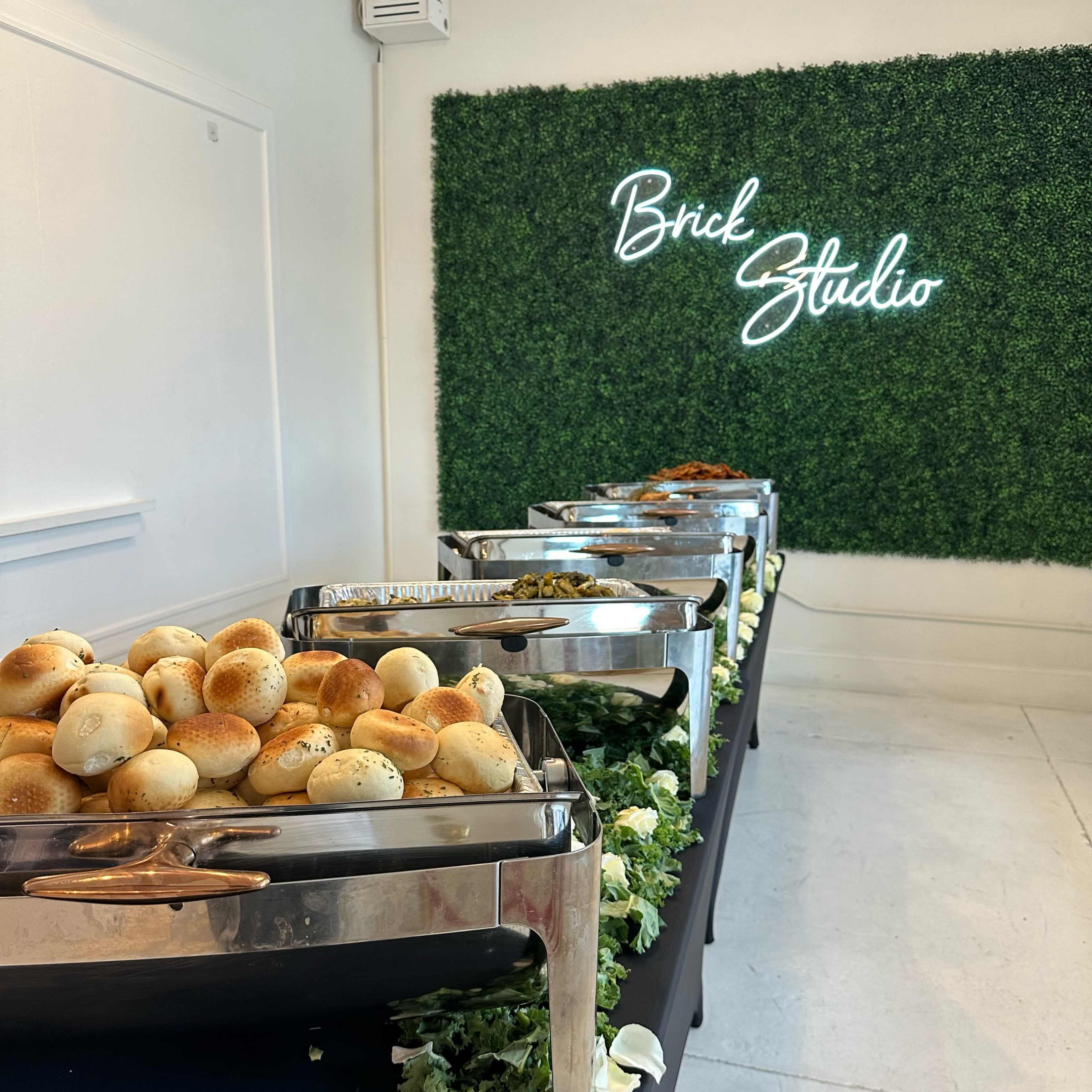 A buffet table displaying various food trays, with rolls arranged in a large, stainless steel container, set against a green wall with the words "Brick Studio" illuminated in neon.