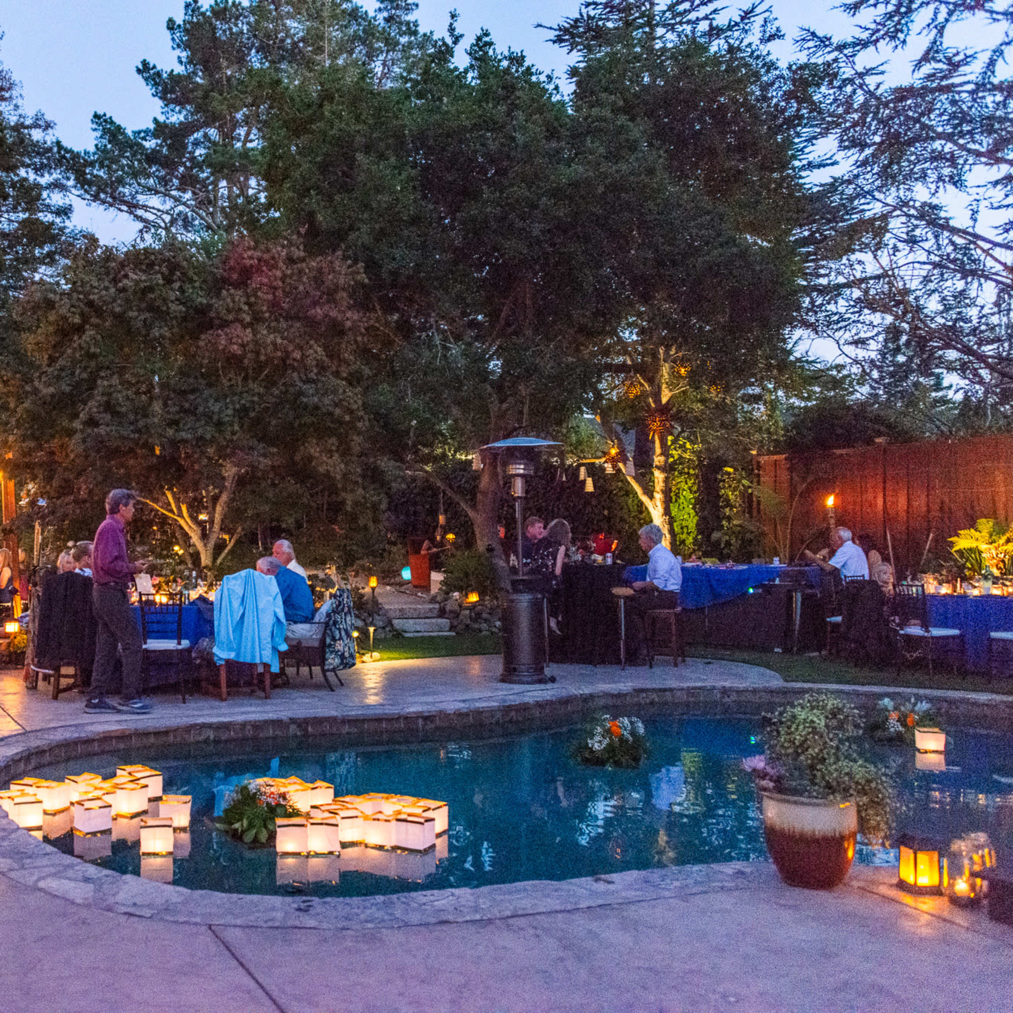 An outdoor event takes place in a garden setting with tables arranged around a pool, illuminated by string lights and floating lanterns.