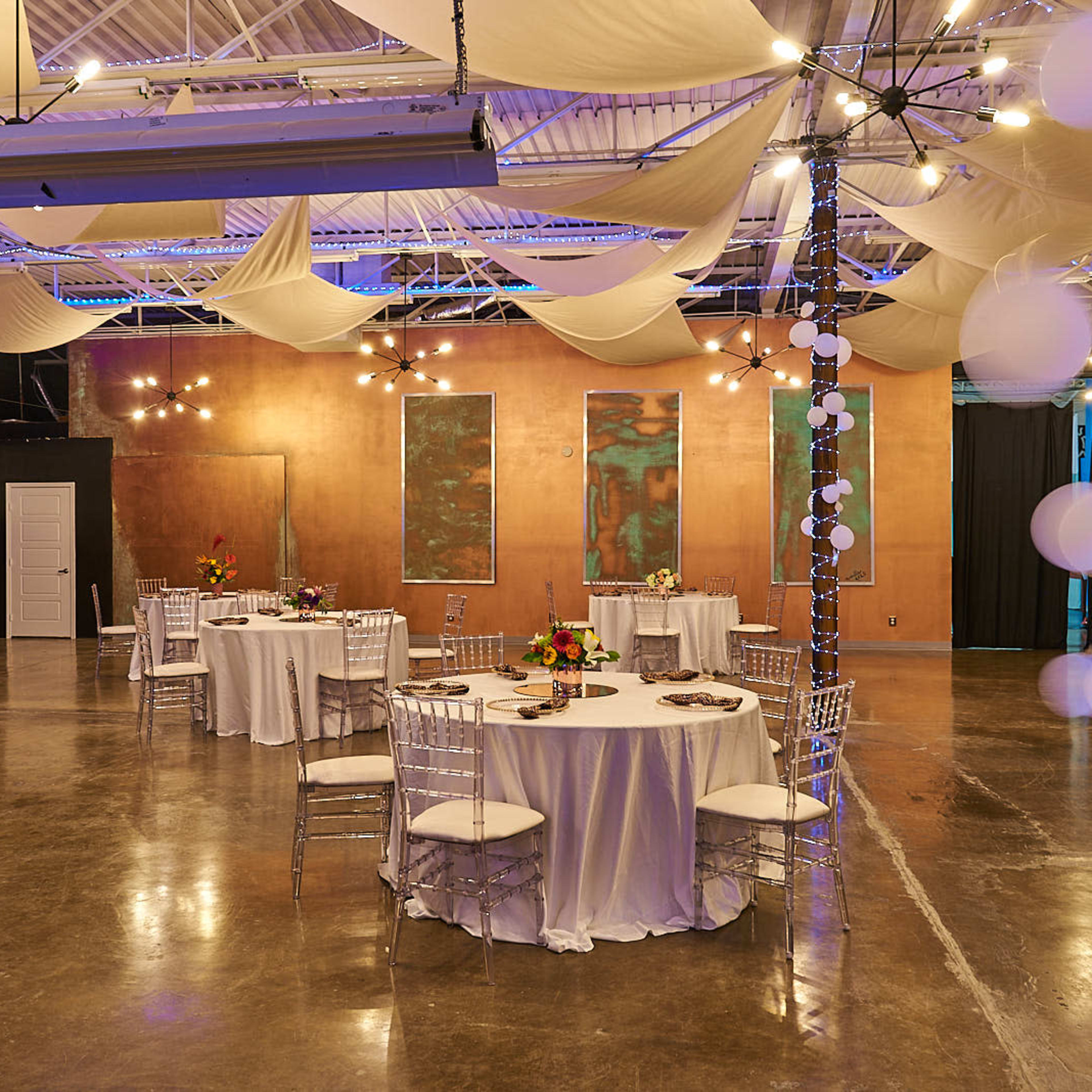An event space features round tables with white tablecloths and floral centerpieces, surrounded by draped fabric and string lights.