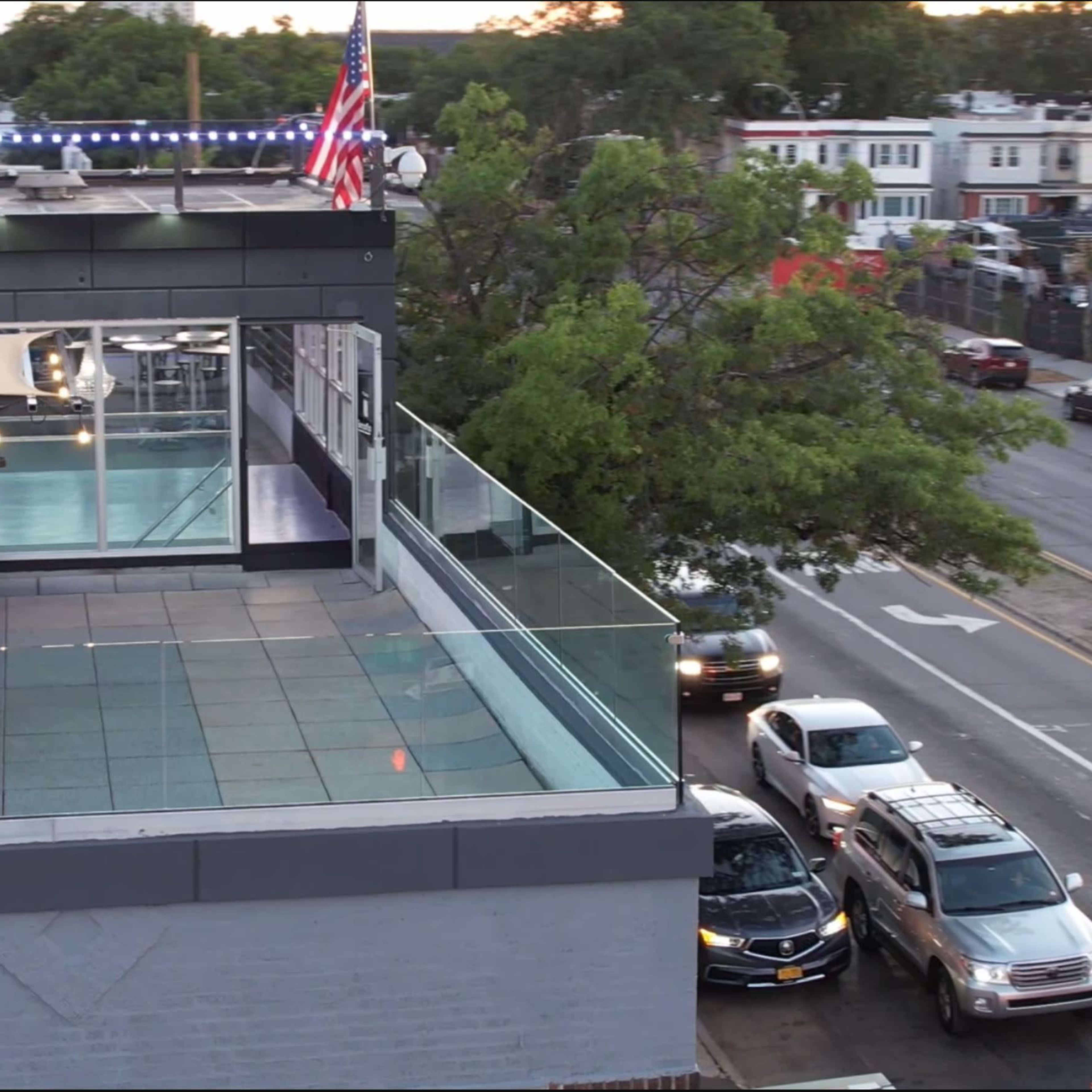 A rooftop terrace with an American flag, overlooking a street lined with parked cars and buildings in the background.