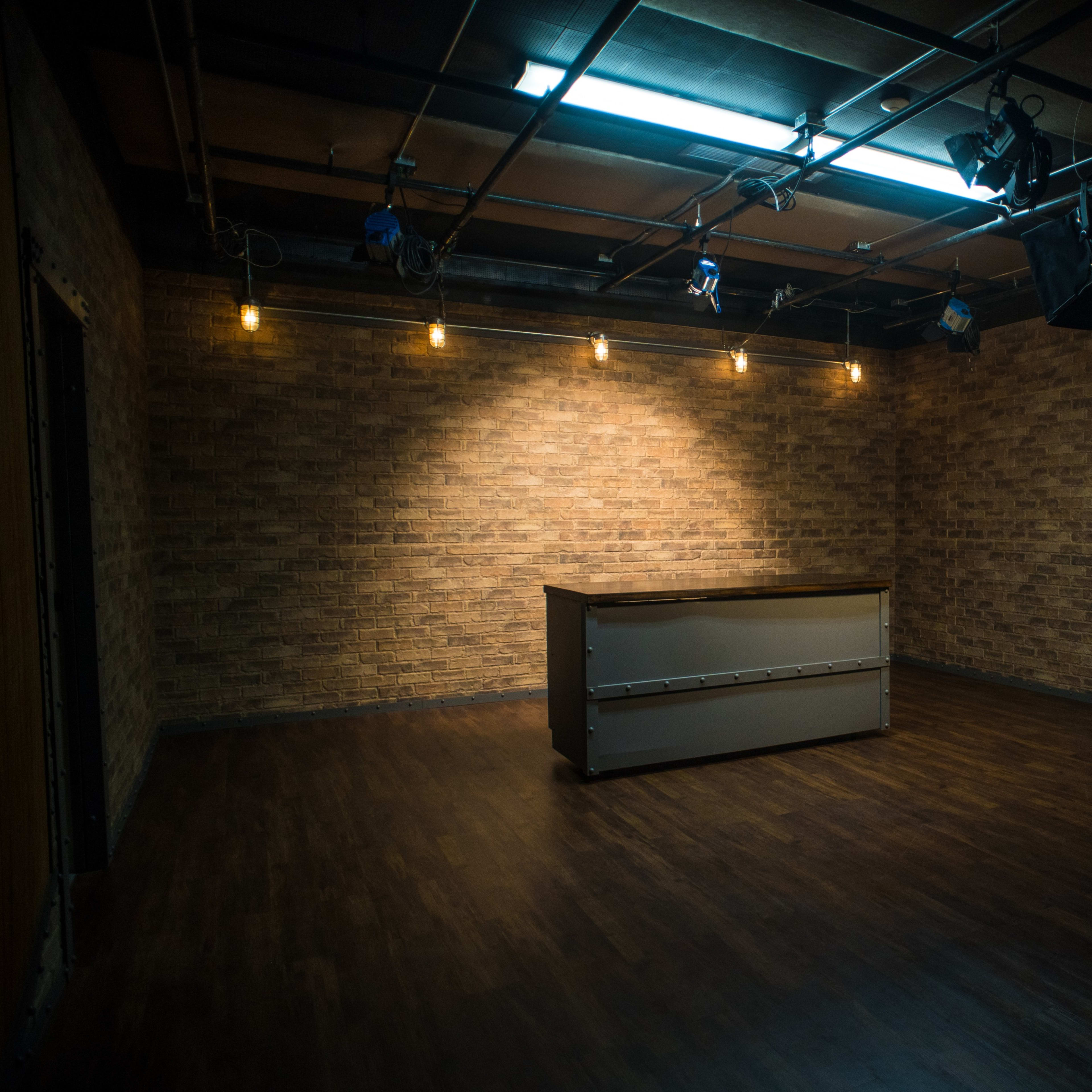An empty studio space with a wooden floor, exposed brick walls, and a central counter lit by overhead studio lights.