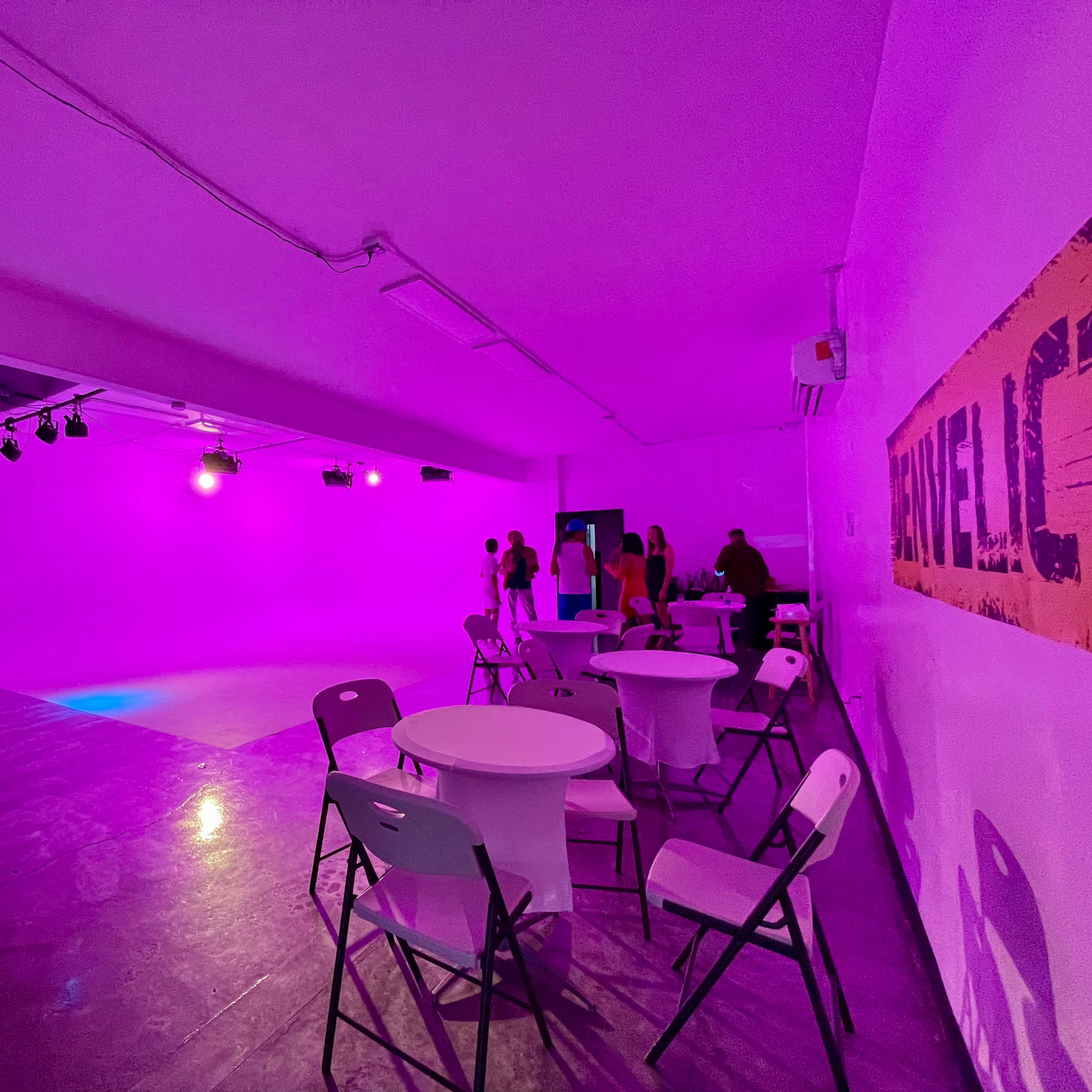 A brightly lit, purple-themed event space with several white tables and chairs, along with a small DJ setup in one corner.