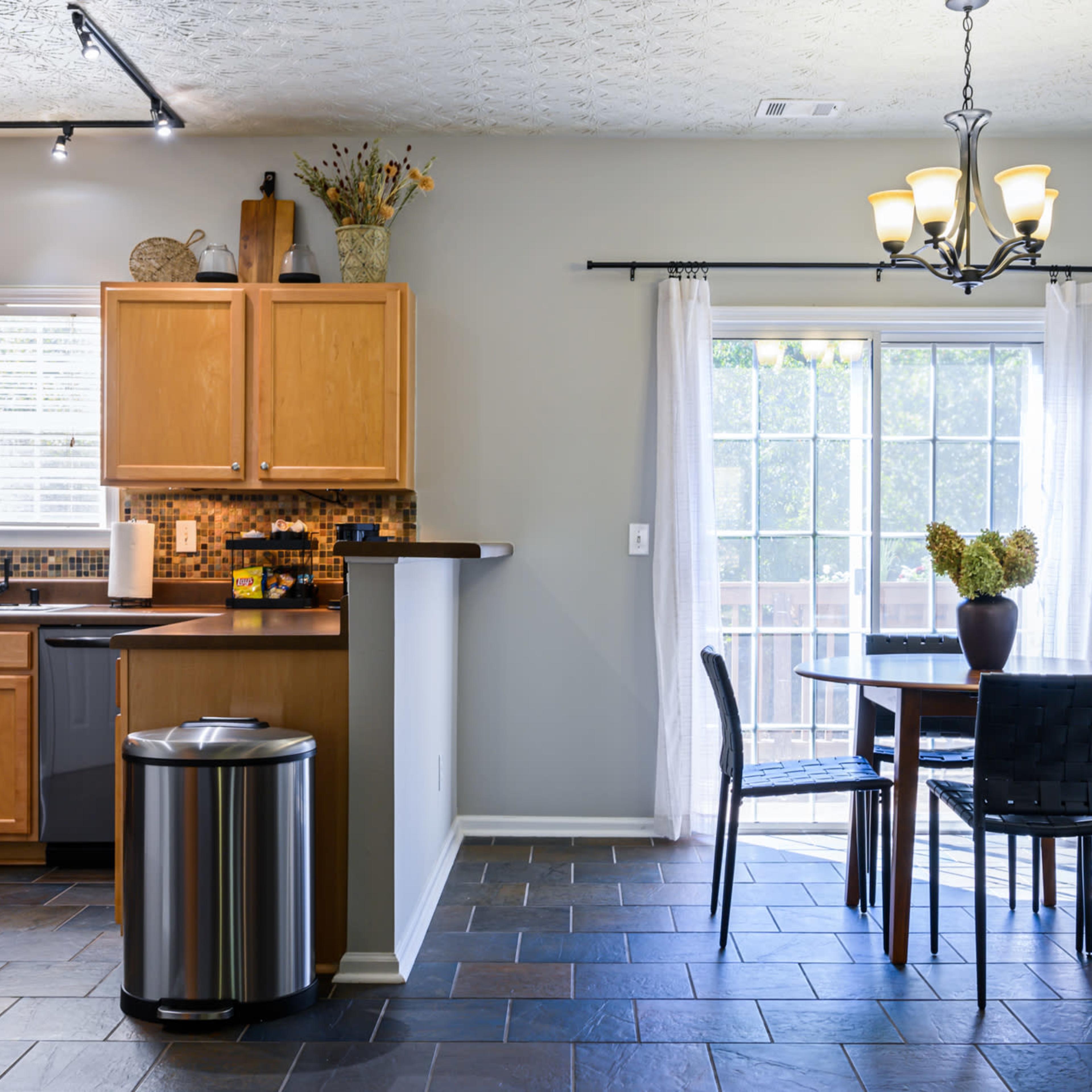 A modern kitchen and dining area with wooden cabinets, a round table set for four, and a large window leading to a patio.