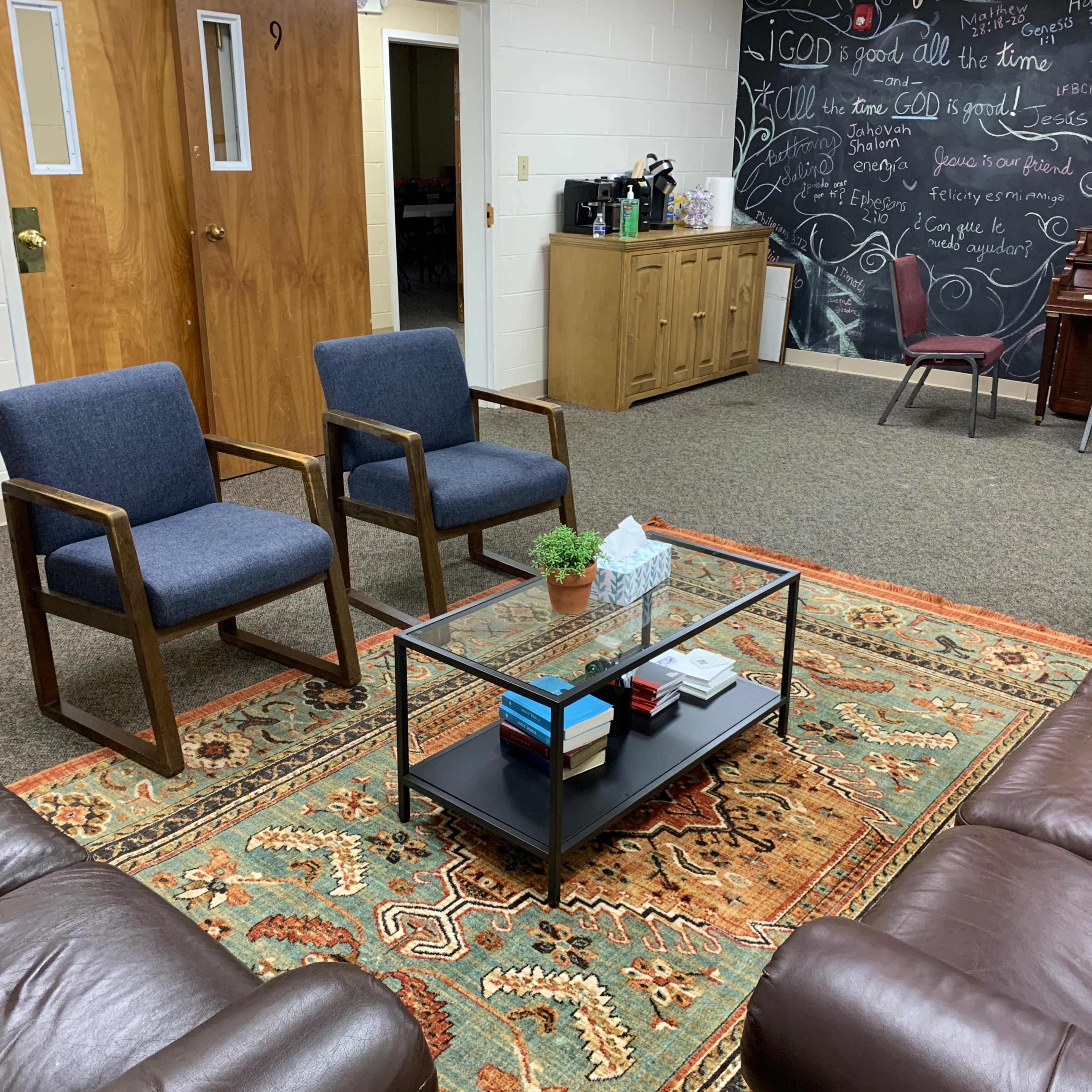 A sitting area with two upholstered chairs facing a coffee table on a patterned rug, surrounded by a couch and a chalkboard wall in the background.