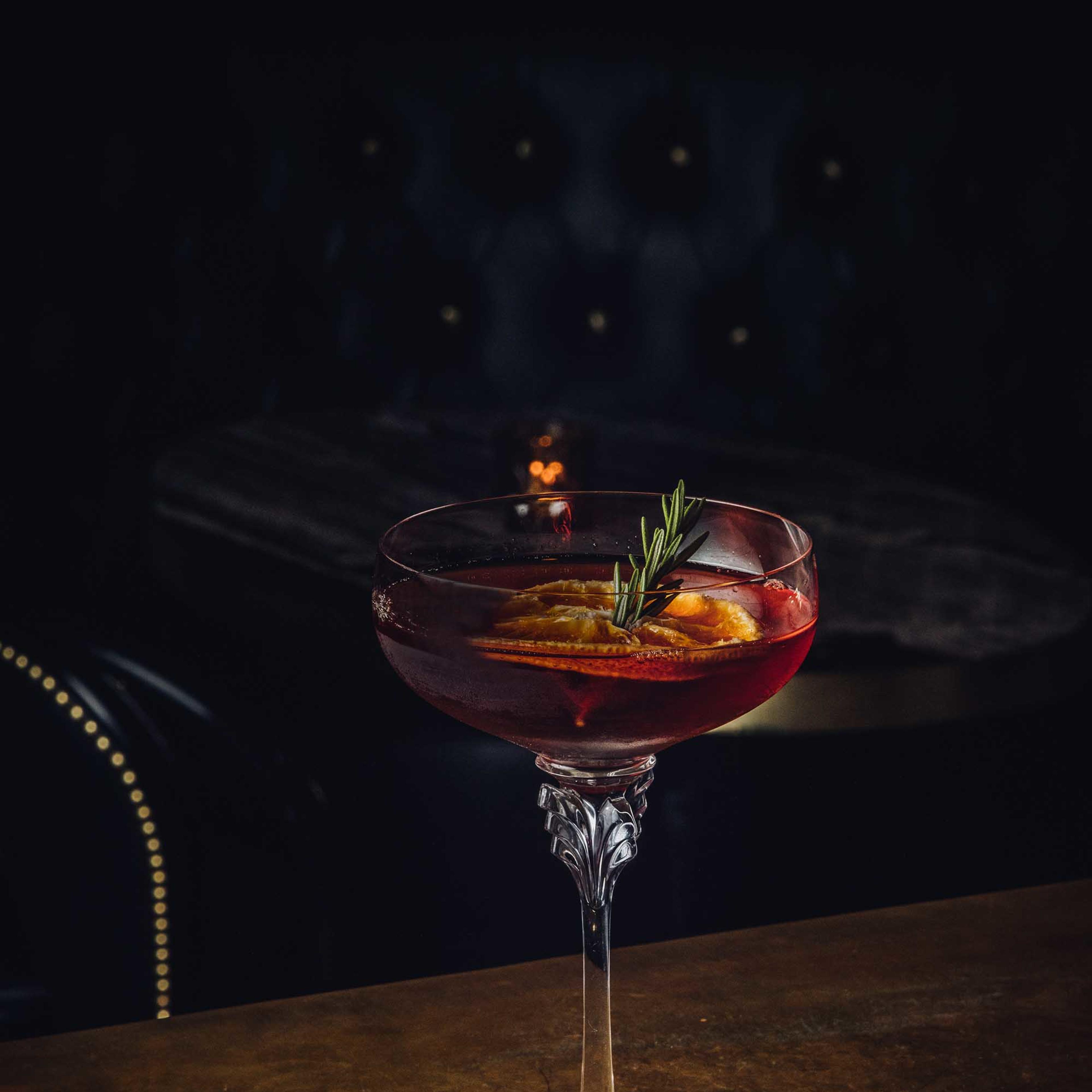 A cocktail garnished with orange slices and a sprig of rosemary sits on a reflective gold table under dim lighting.
