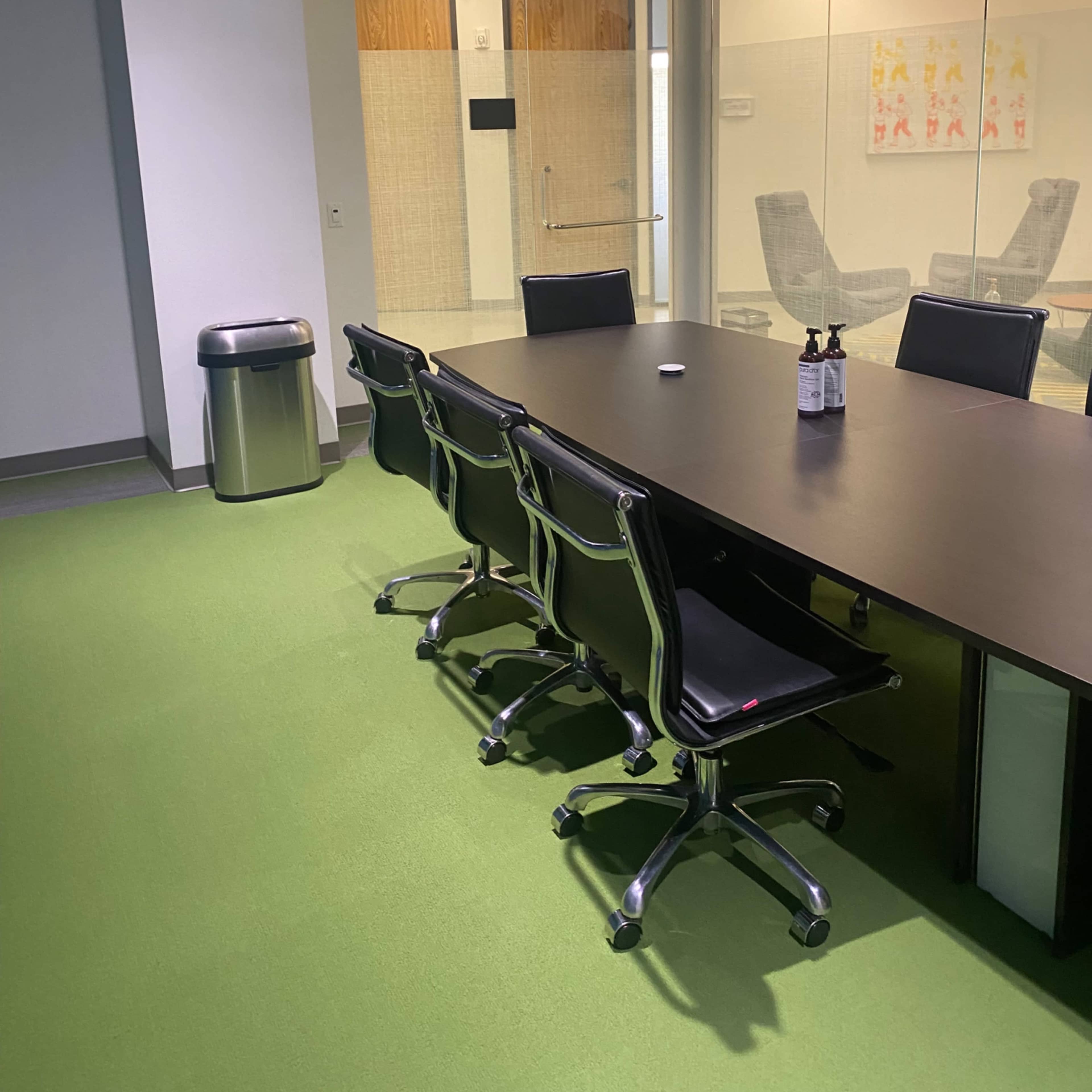 A conference room featuring a large, dark table surrounded by black rolling chairs, with green carpet and a glass-walled meeting space visible in the background.