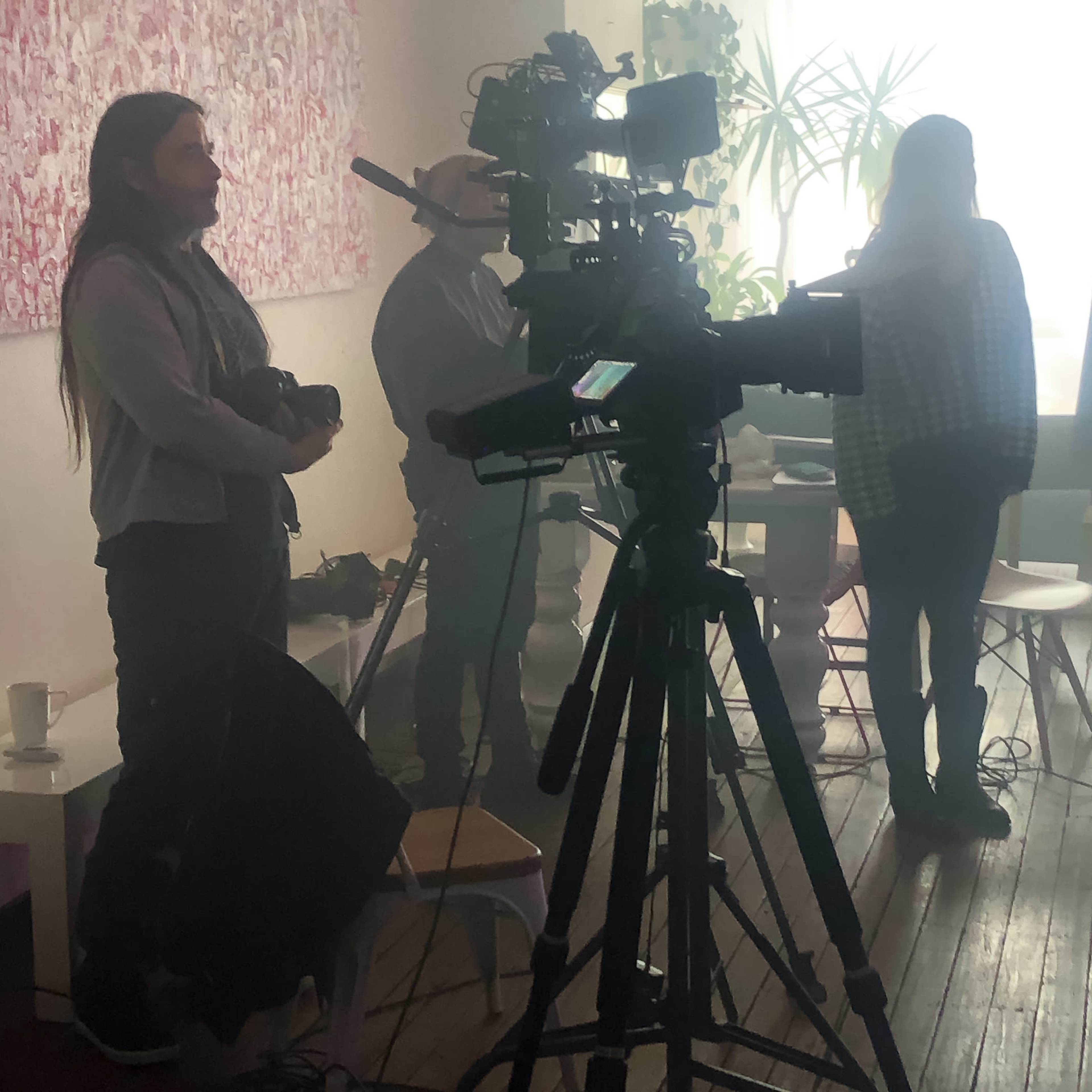A diverse group of people stands behind several cameras in a bright, airy room with plants and art on the walls.