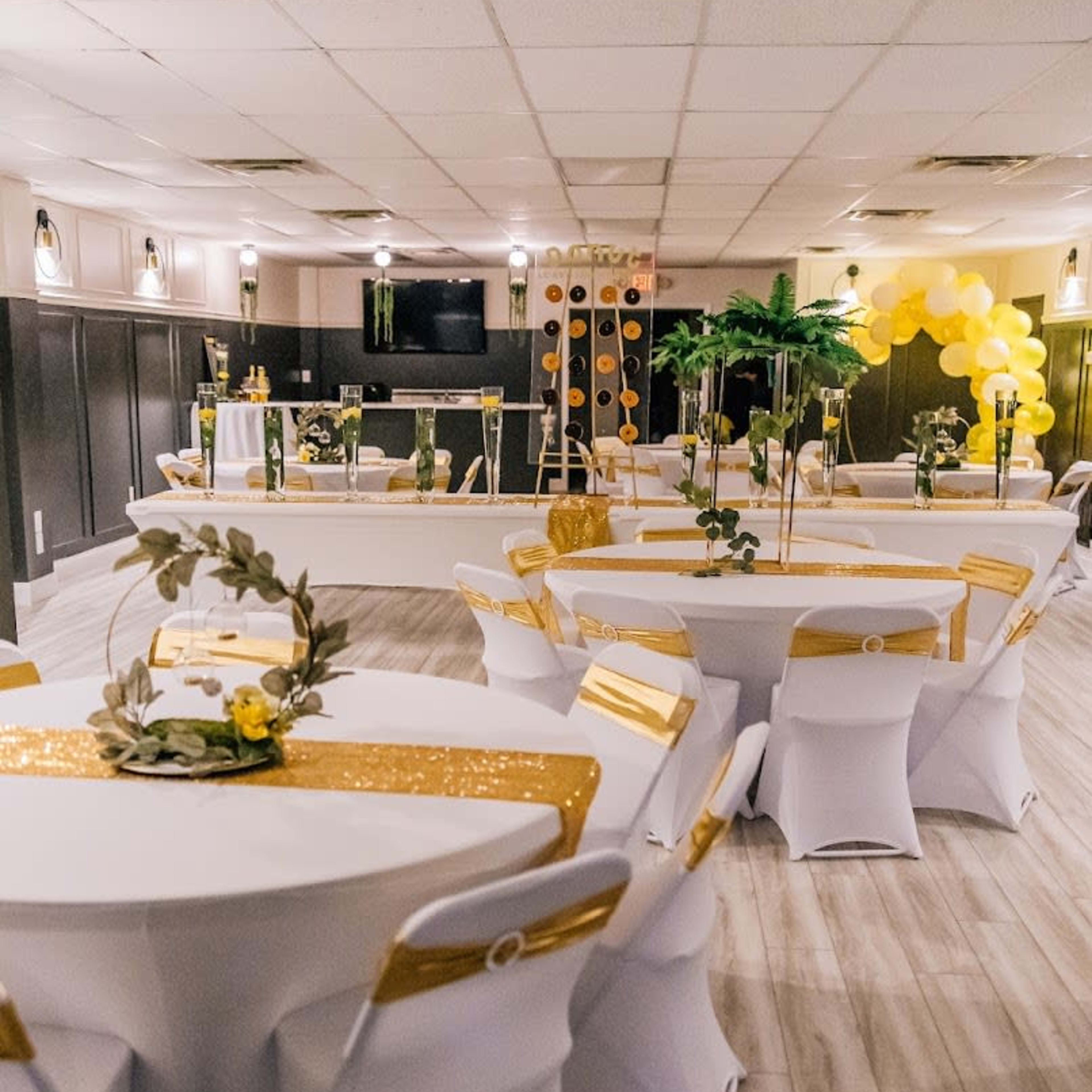 A banquet hall is set up with round tables covered in white linens and gold accents, featuring decorations of greenery and yellow flowers.
