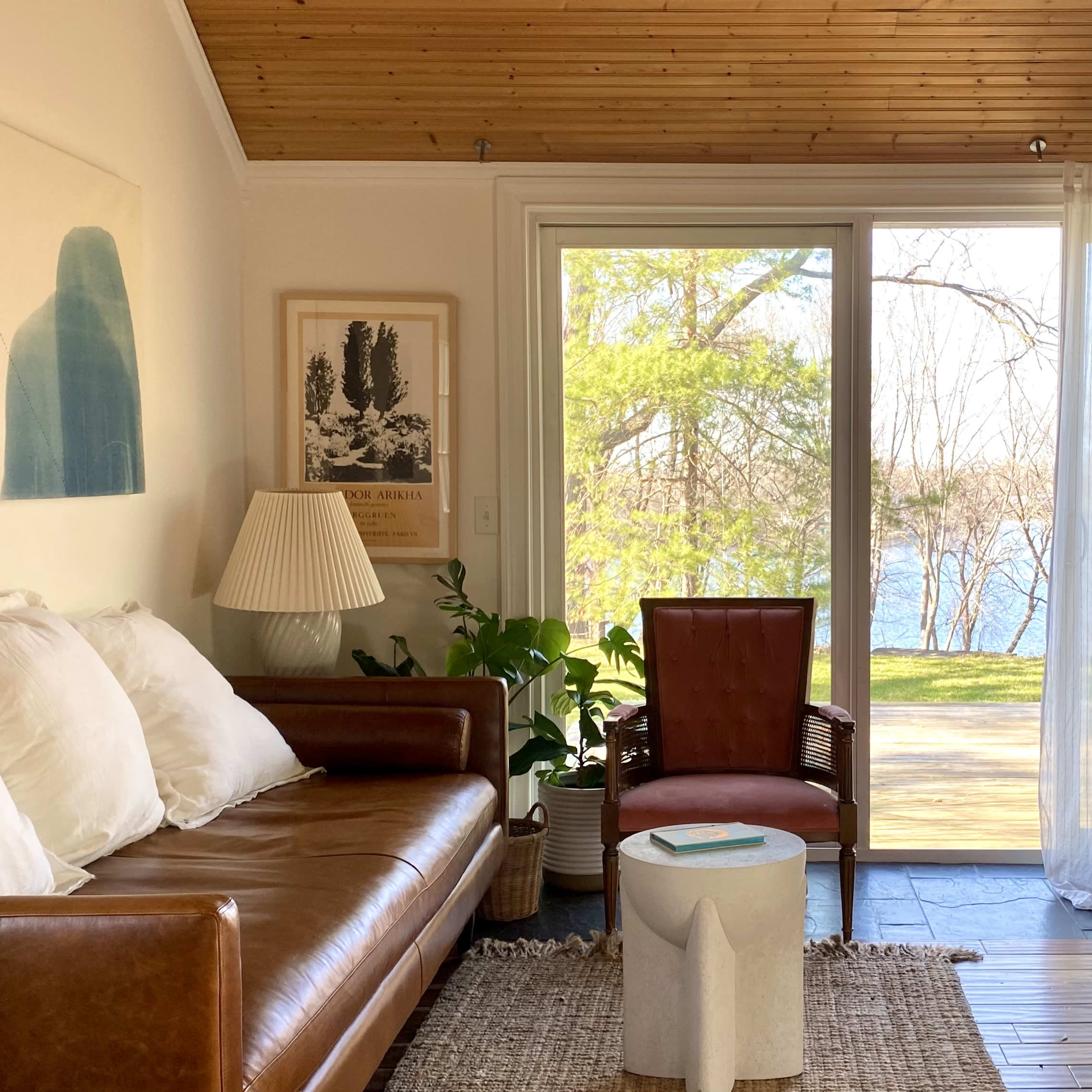 A cozy living room features a brown leather sofa, a wooden chair, a round white table, and large windows with sheer curtains overlooking a lake.