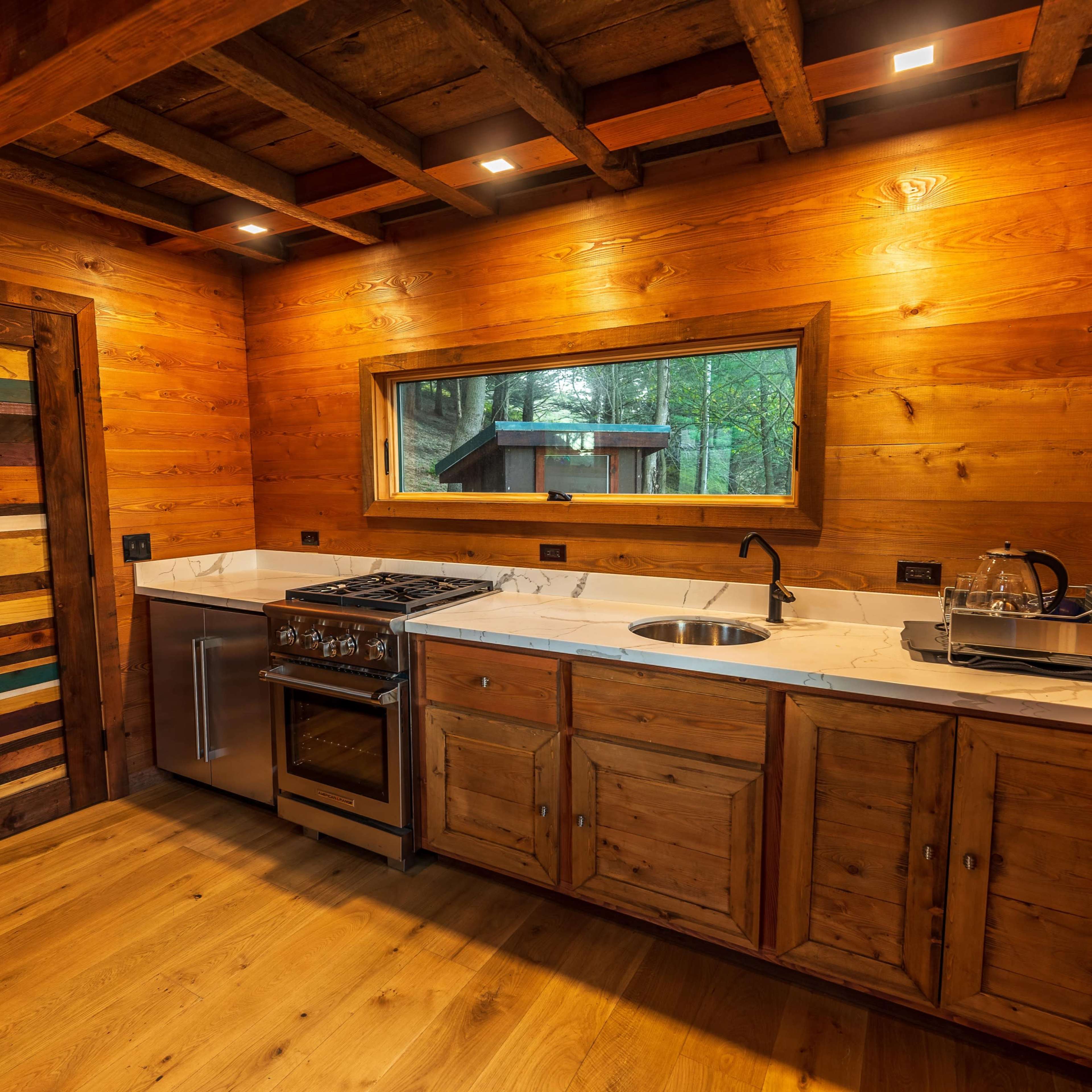 A rustic kitchen with wooden walls, a stove, a sink, and a large window that offers a view to the outdoors.