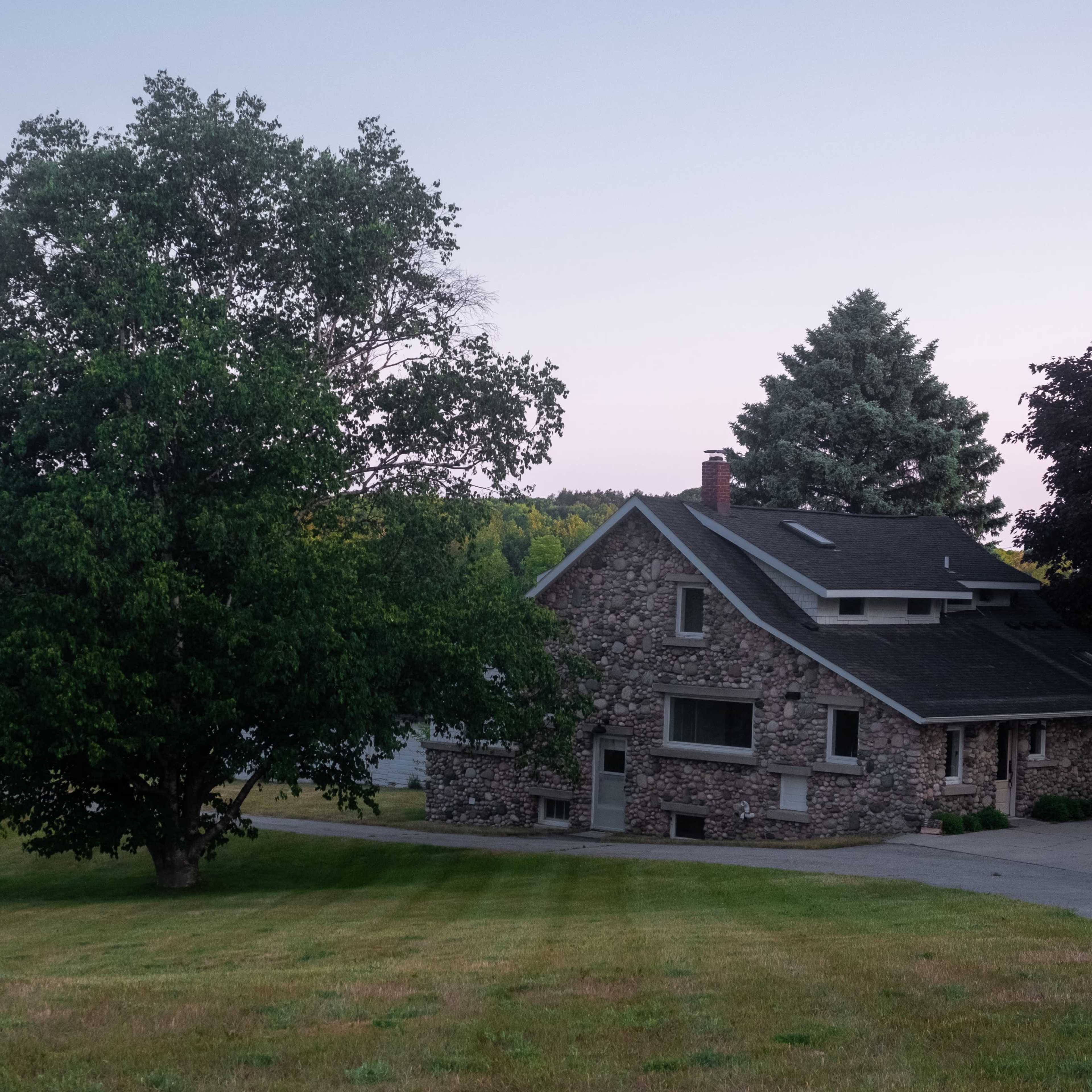 A stone house with a sloped roof is situated near a large tree on a grassy lawn.