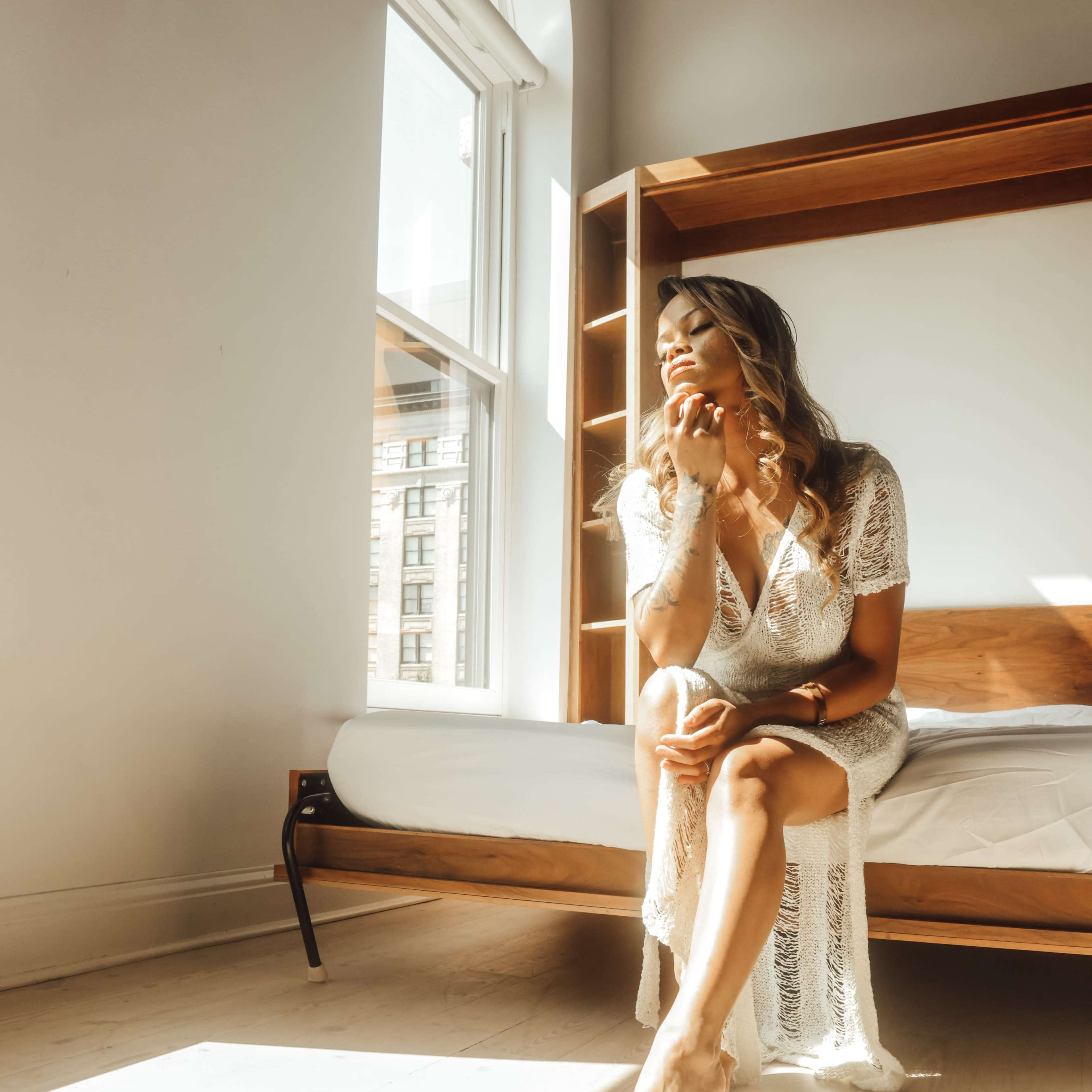 A woman in a lace robe sits on the edge of a bed in a sunlit room with large windows.