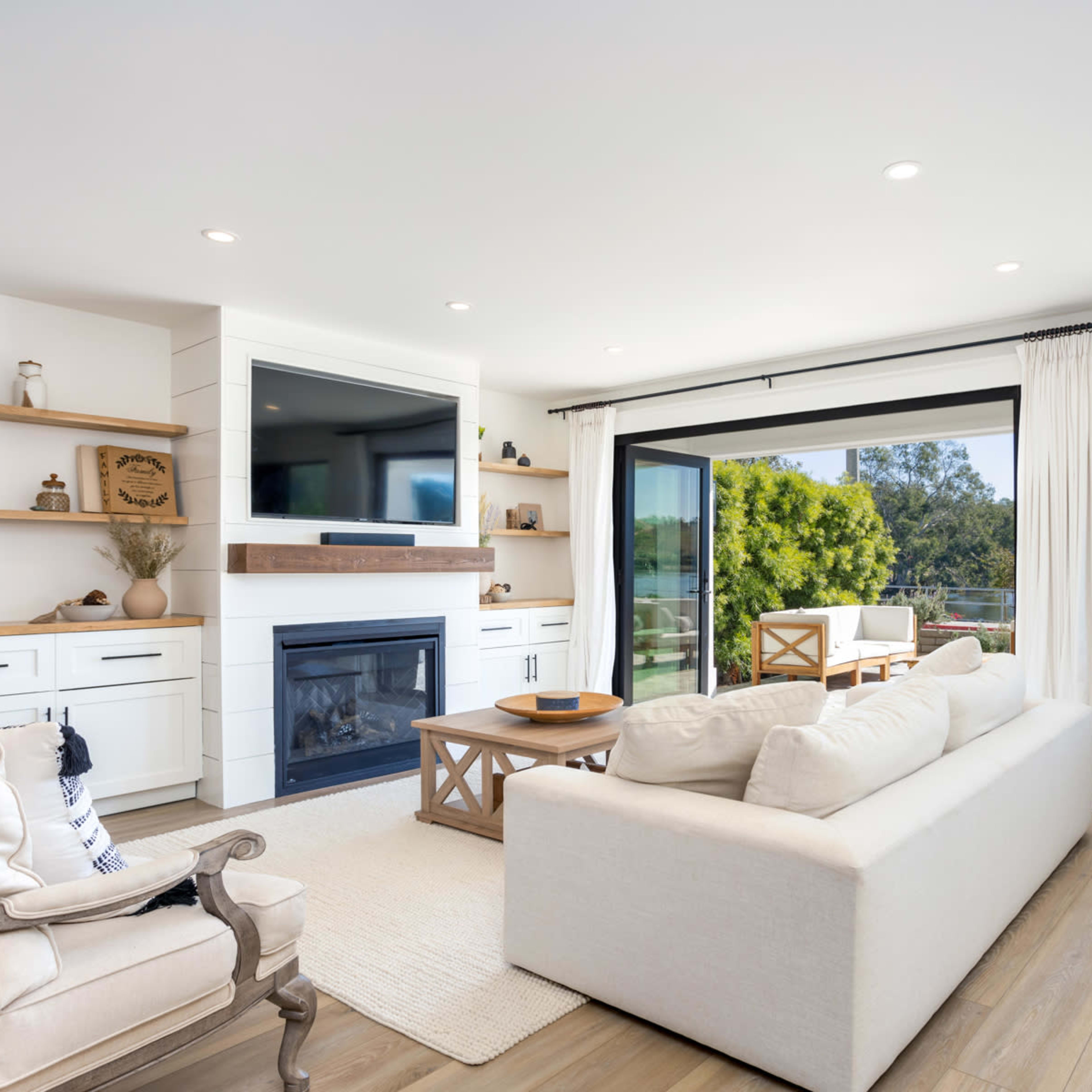 A modern living room features a white sofa, a wooden coffee table, and a television mounted on a light-colored wall, with large windows overlooking a scenic view.