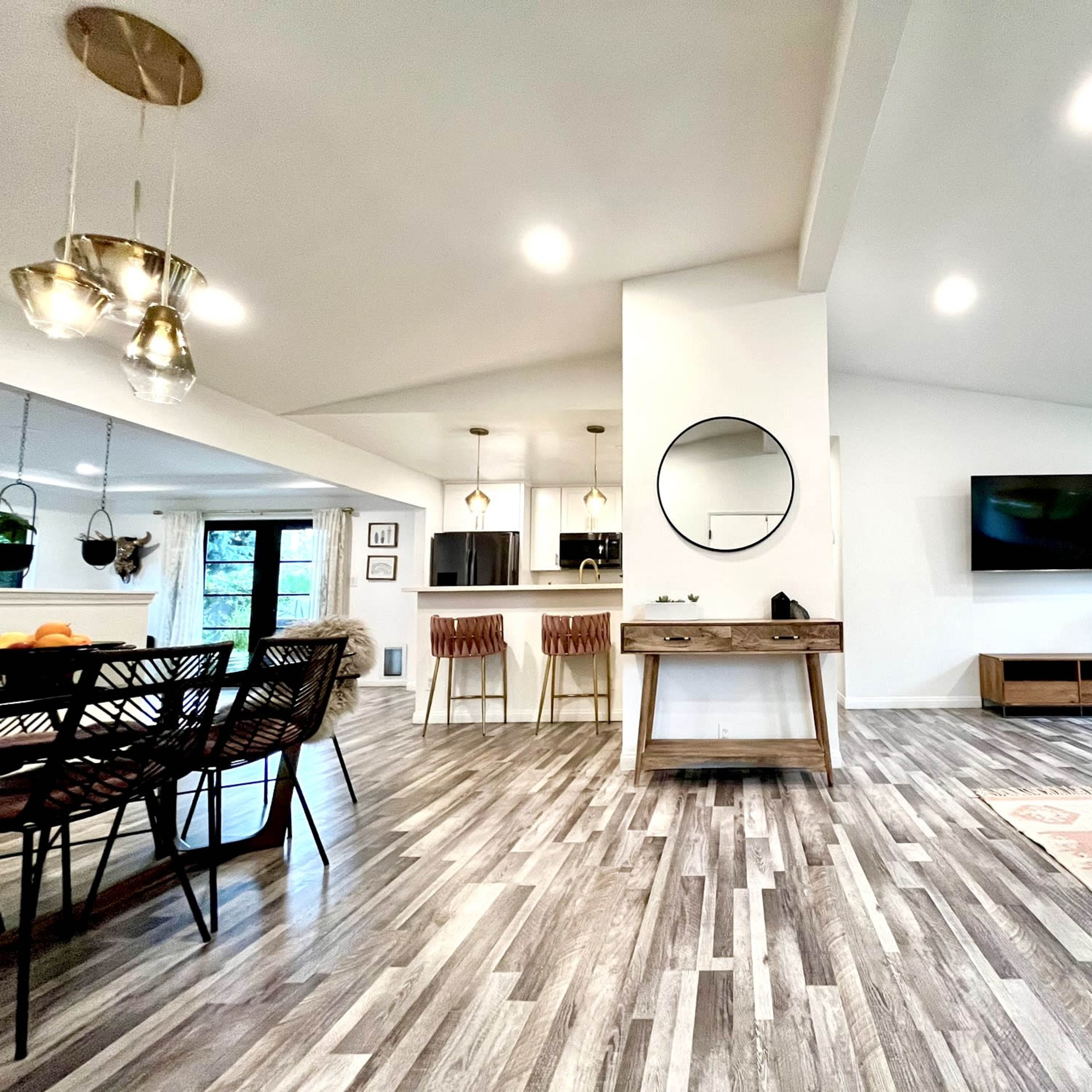 An open-concept living and dining area with wooden flooring, a circular mirror on the wall, and large windows allowing natural light to enter.