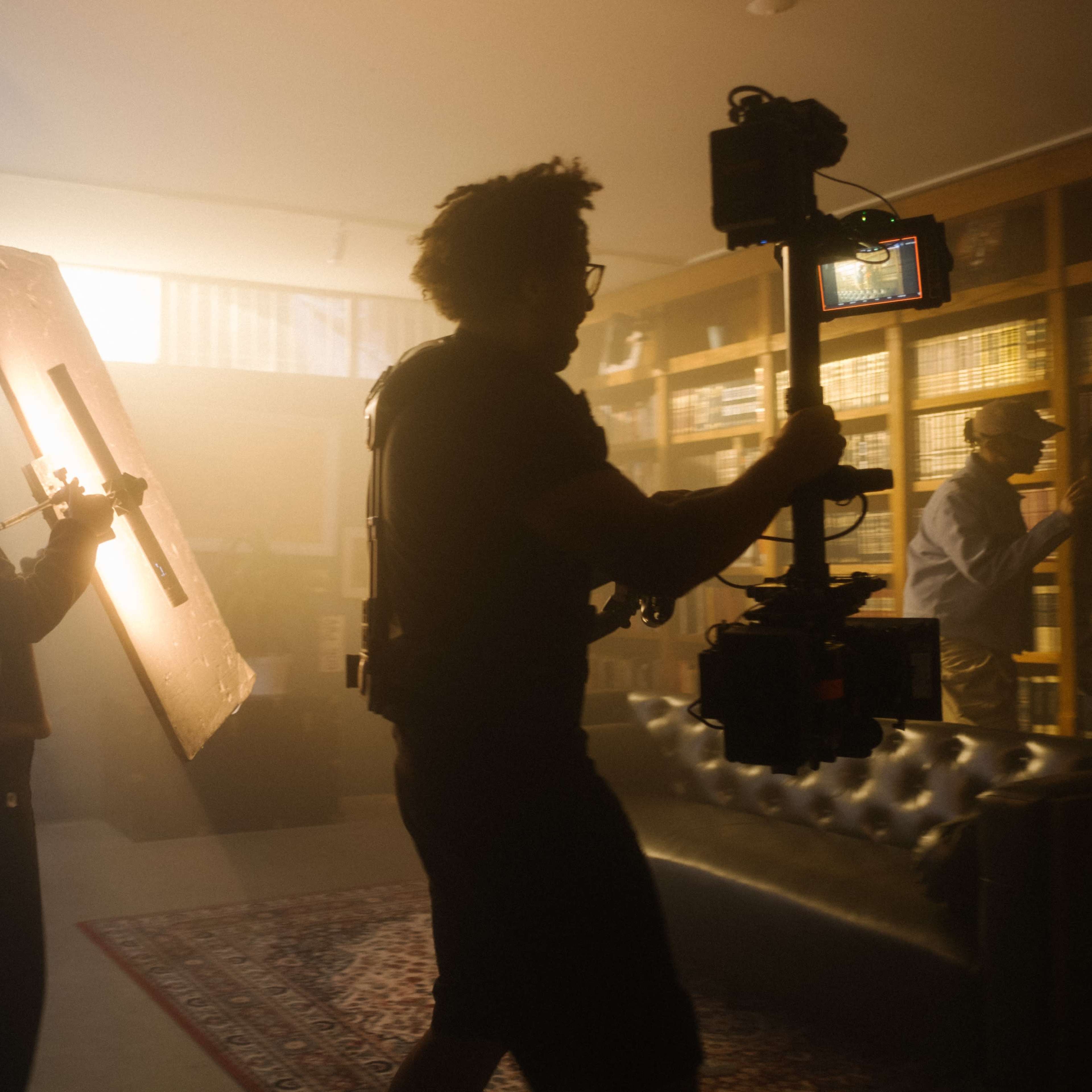 A crew member holds a reflector while another operates a camera in a dimly lit room filled with shelves of books.