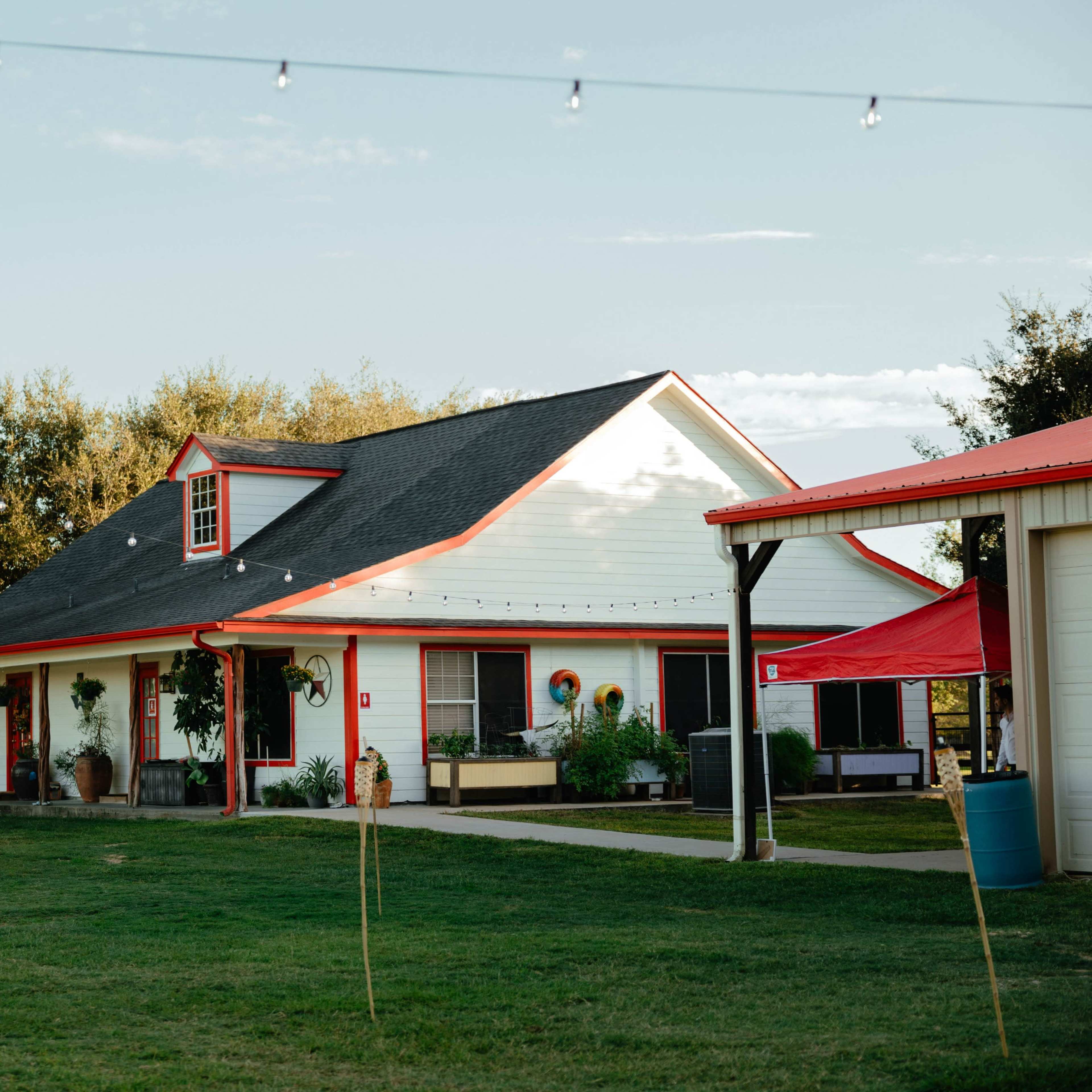 A white house with red accents sits in a grassy area, surrounded by trees and string lights.